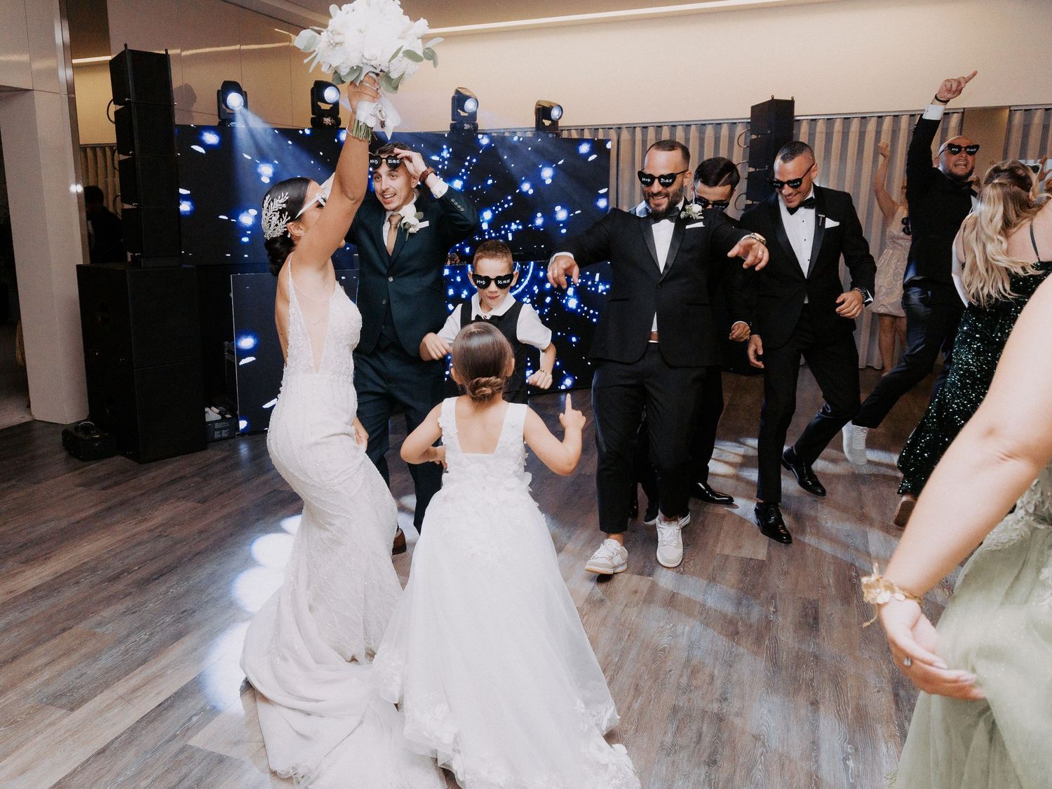 Joyful wedding party dancing on wooden floor during reception celebration in elegant venue.