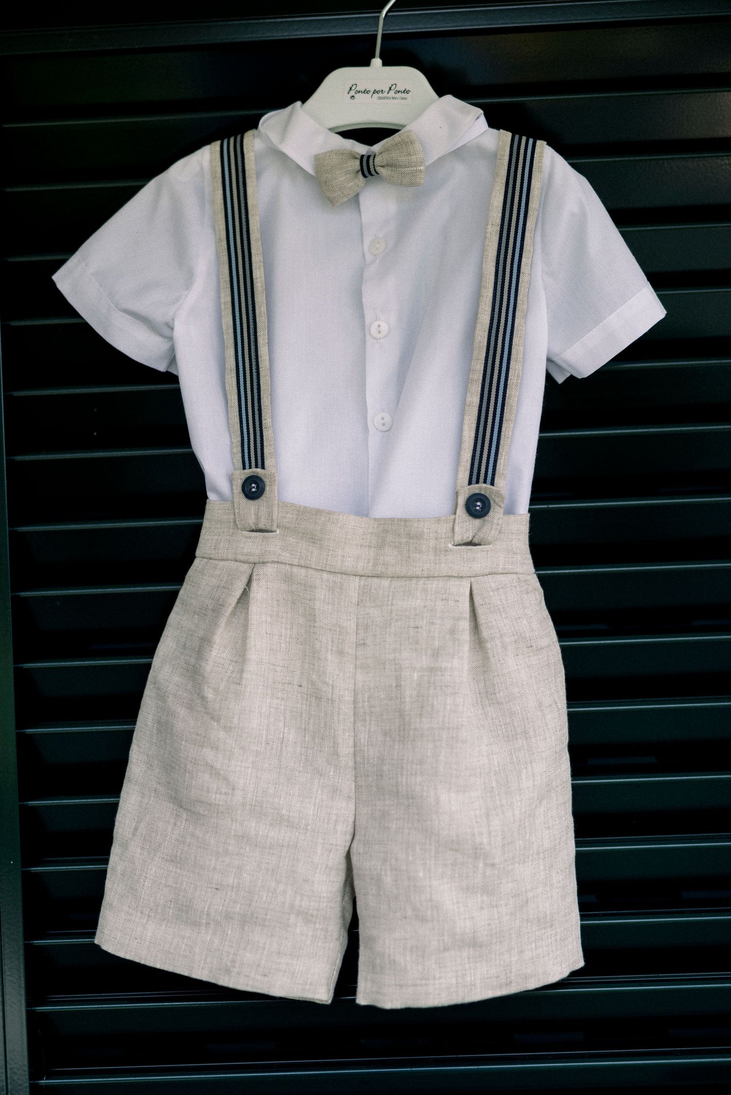 Conjunto formal para bebê, composto por camisa branca, bermuda de linho bege e suspensórios listrados azul-marinho, em exposição. Batizado