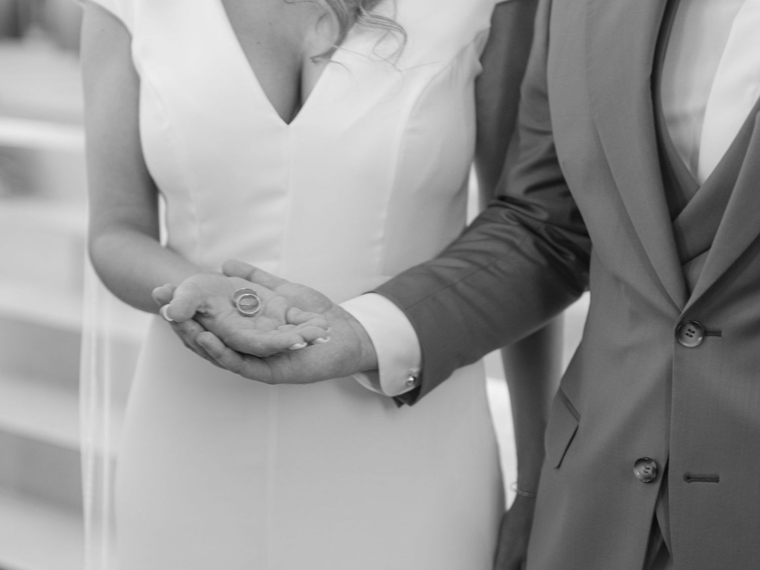 A close-up black and white photo of a couple holding hands, showing wedding attire and rings.