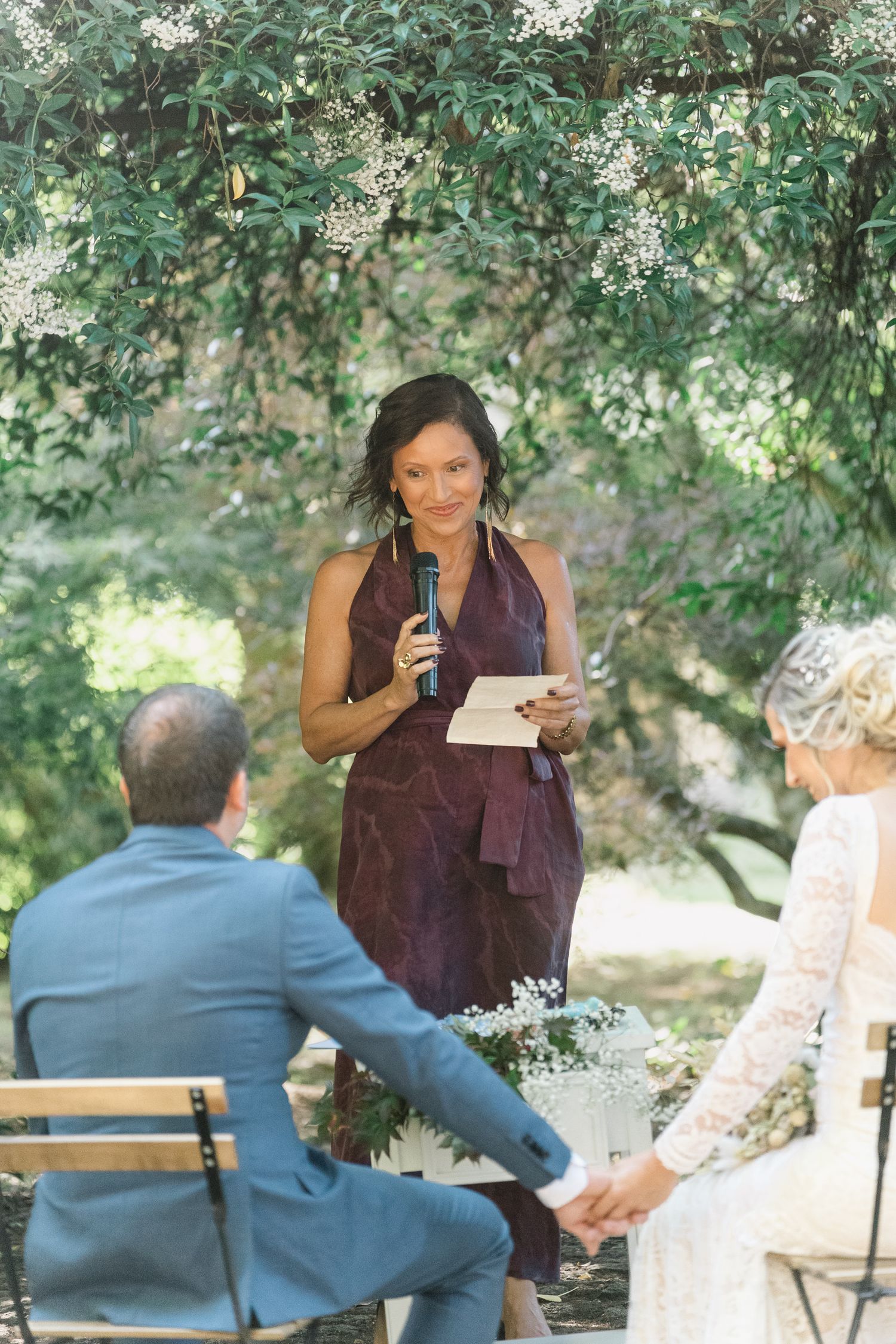 A person in a burgundy top reads from papers while another person in blue sits on a bench under shady trees.