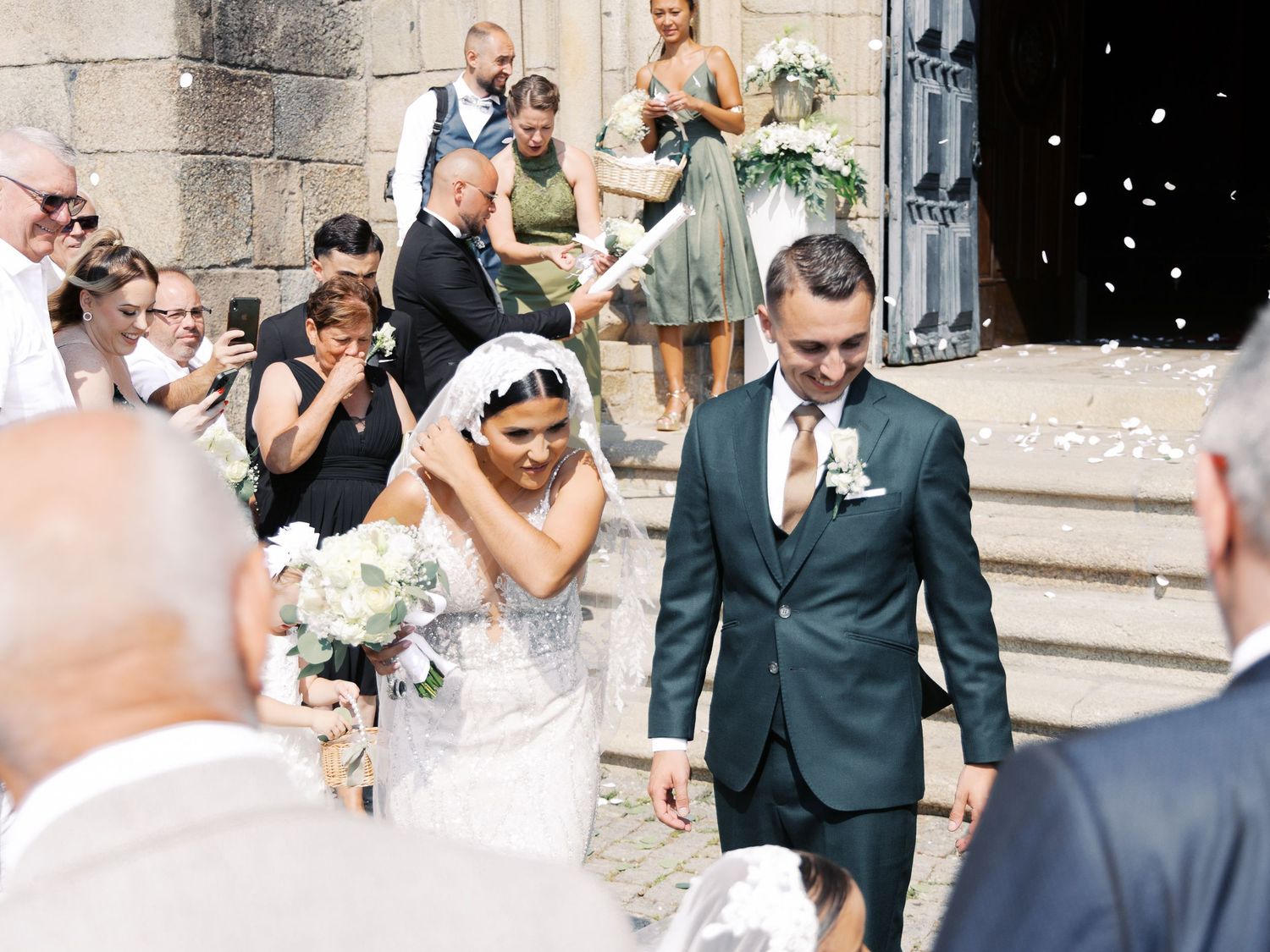Wedding party celebrates with confetti and joyful expressions outside a stone building during a summer celebration.