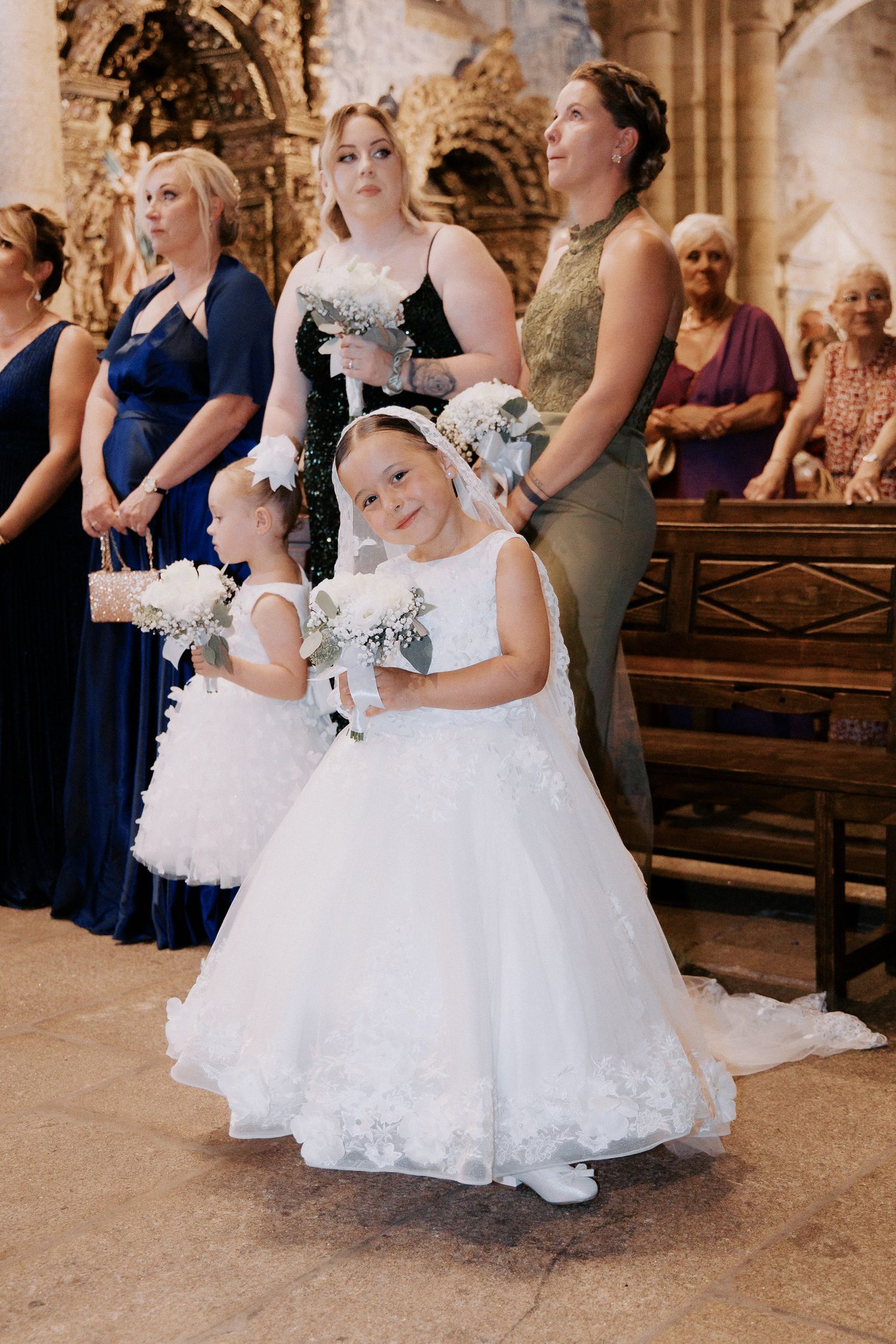 Two young flower girls in white dresses share a sweet moment during a church wedding ceremony.