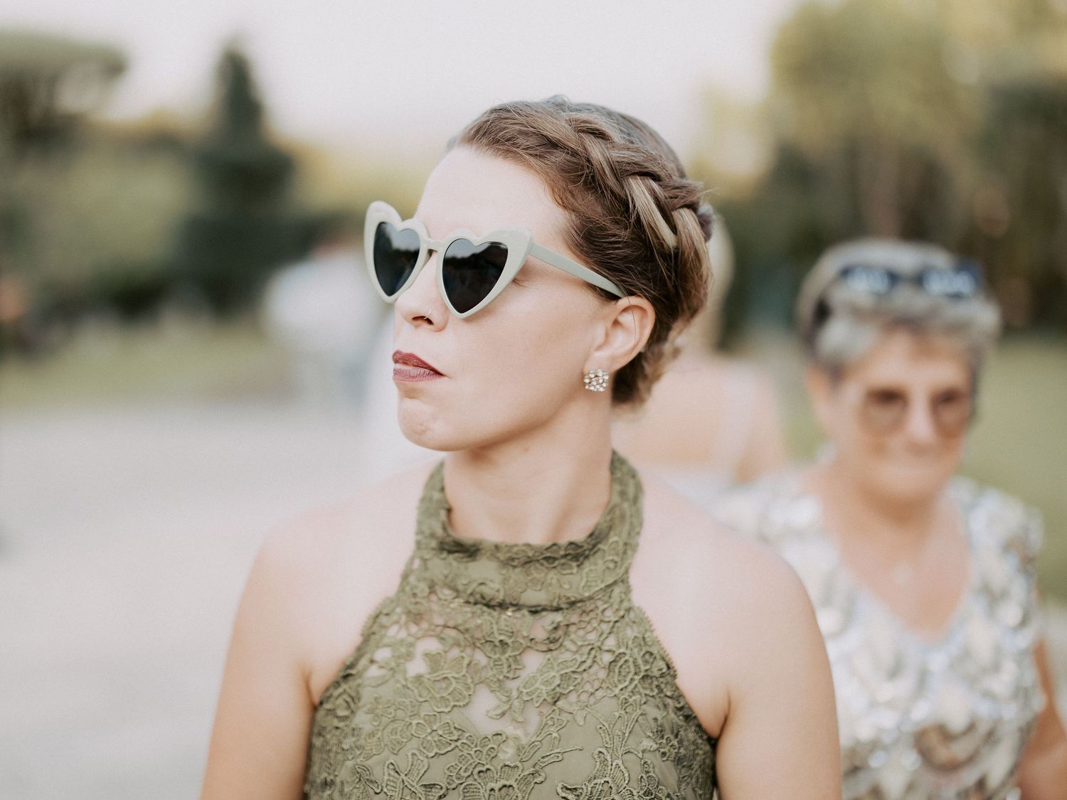 A person wearing heart-shaped sunglasses and a green sequined dress stands outdoors with someone in white behind them.