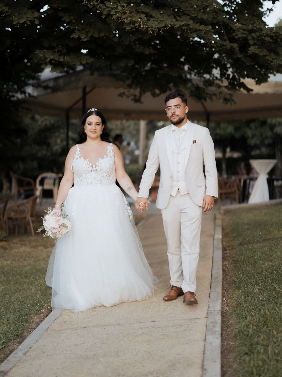 Couple in white wedding attire walking together on grassy outdoor venue with trees in the background.