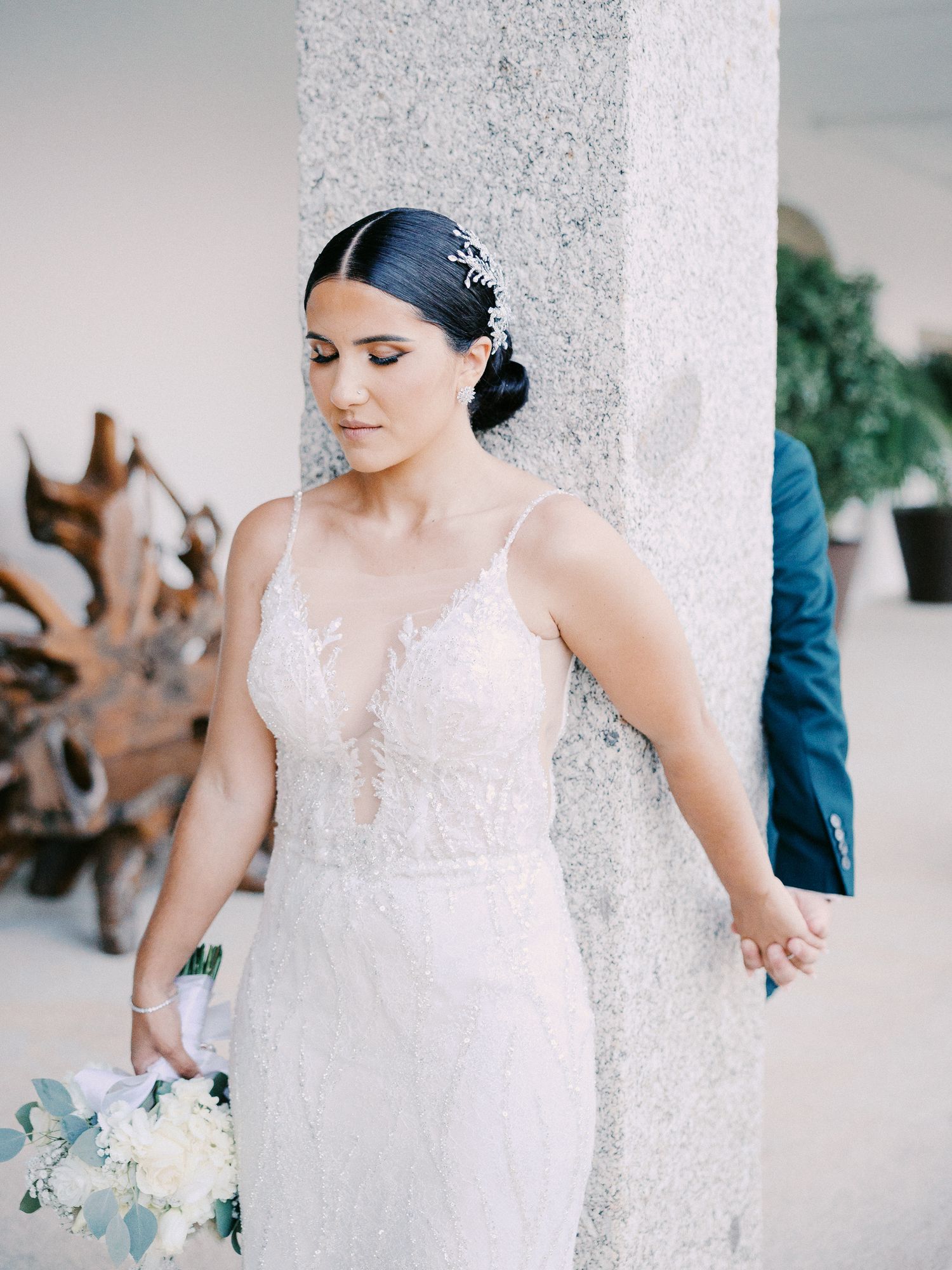 A bride in a white lace wedding dress stands elegantly against a light-colored column.