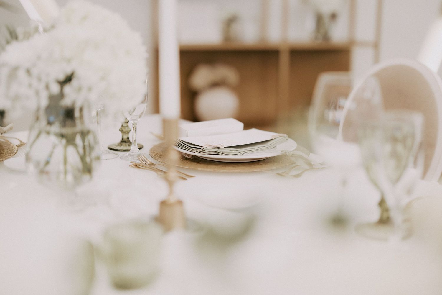 Elegant white table setting with stacked plates and wine glasses in soft focus against a neutral background.