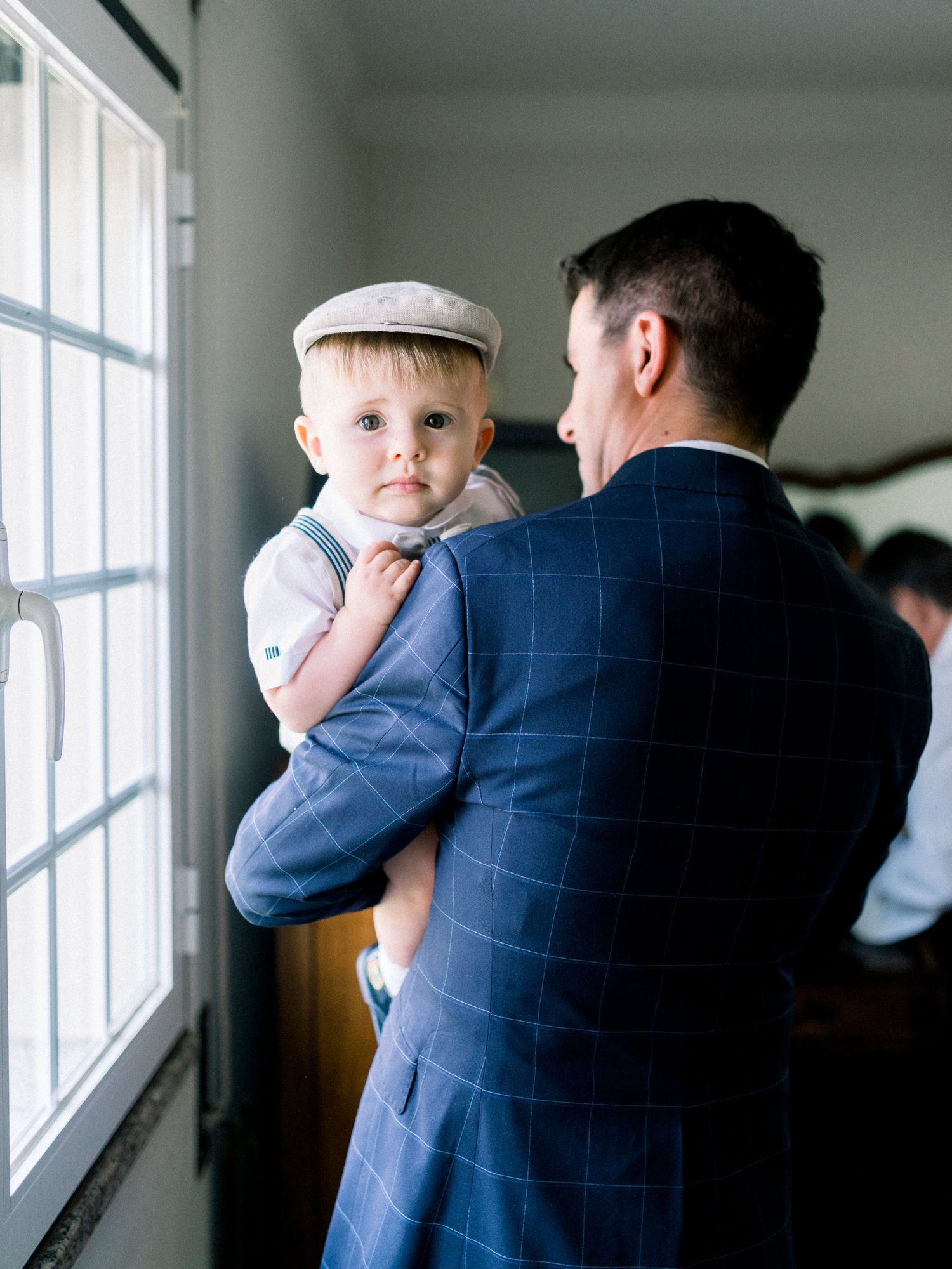 Parent and baby in formal attire gaze out window together in tender moment at home.