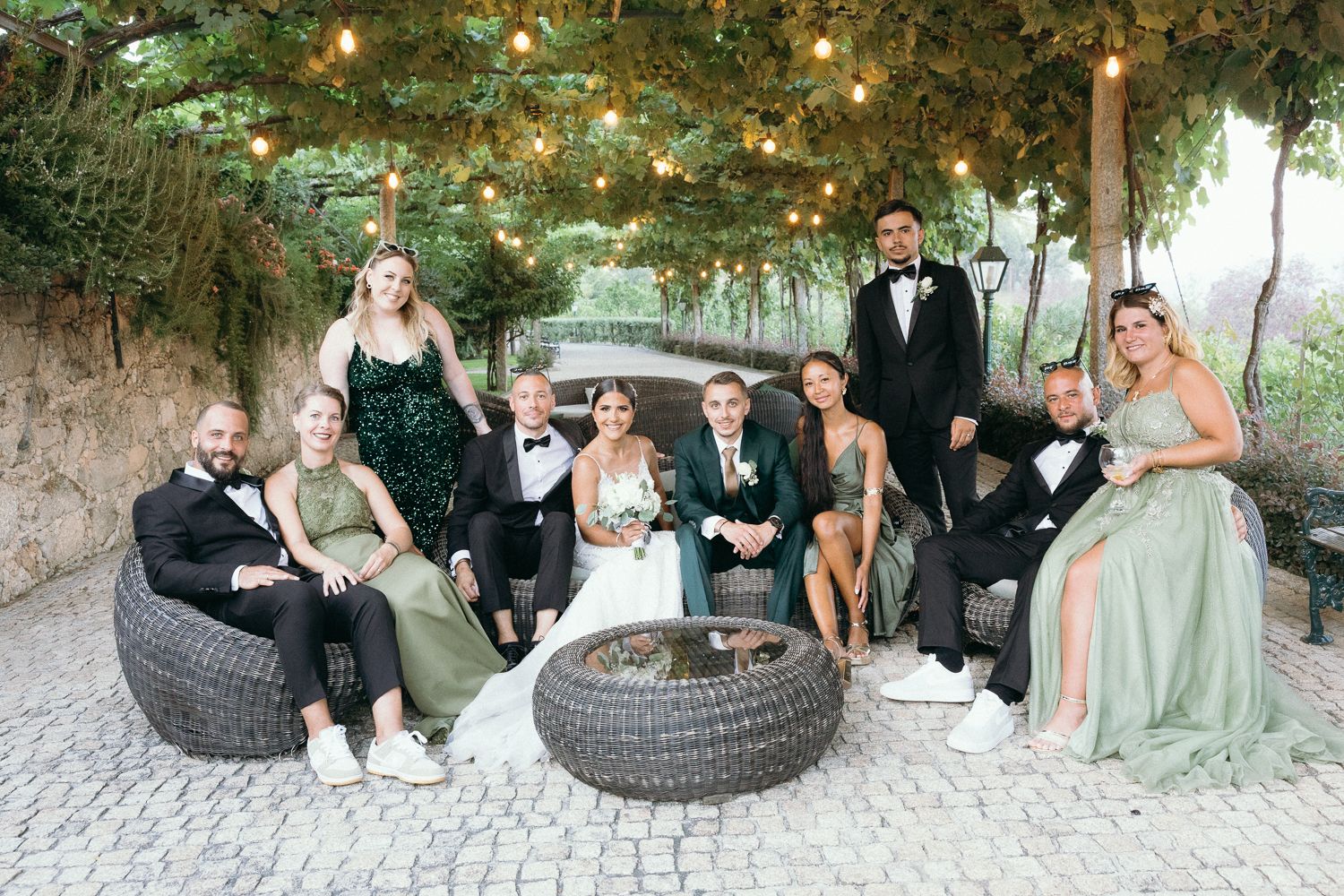 Wedding party relaxes on outdoor patio furniture under string lights and greenery during elegant garden reception.