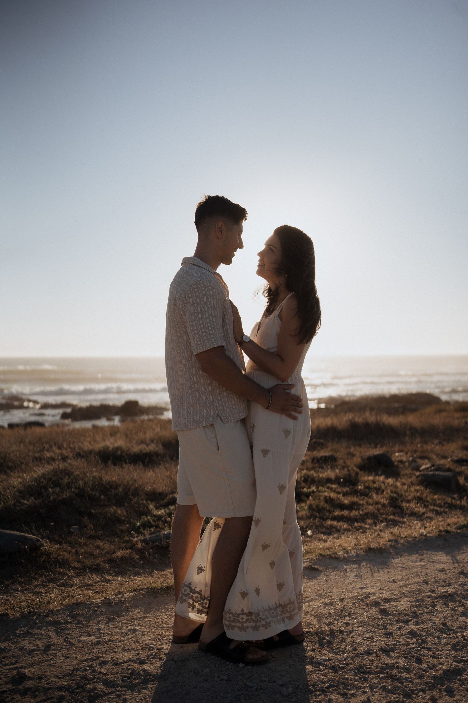 Um casal se abraça em um penhasco costeiro ao pôr do sol, vestindo roupas brancas combinando.