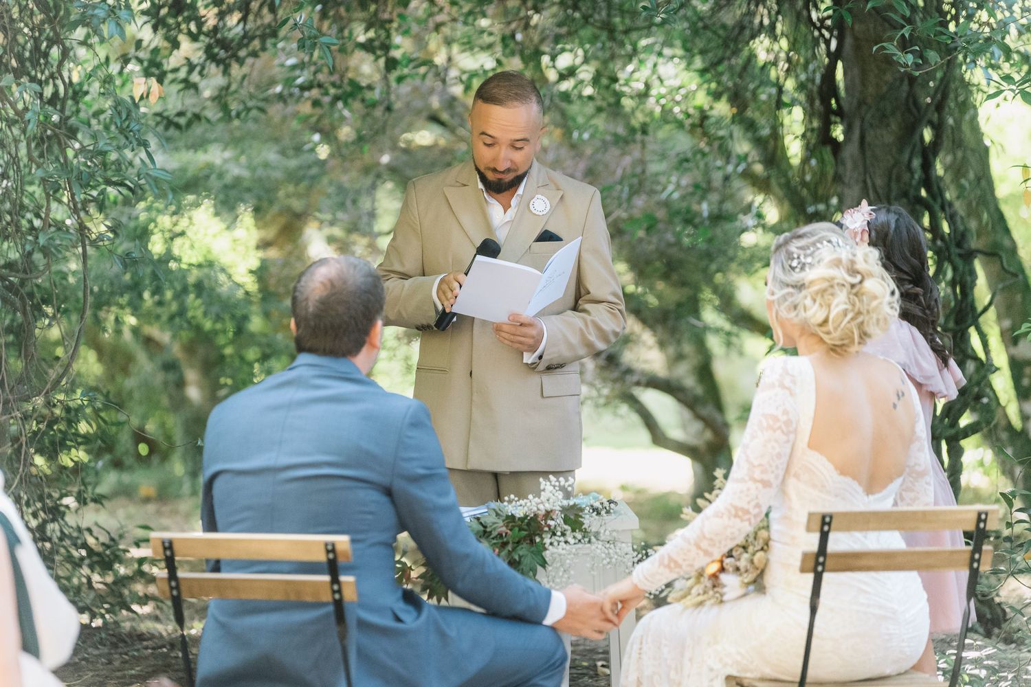A romantic outdoor wedding ceremony takes place in a sunlit garden with wooden chairs arranged on the grass.
