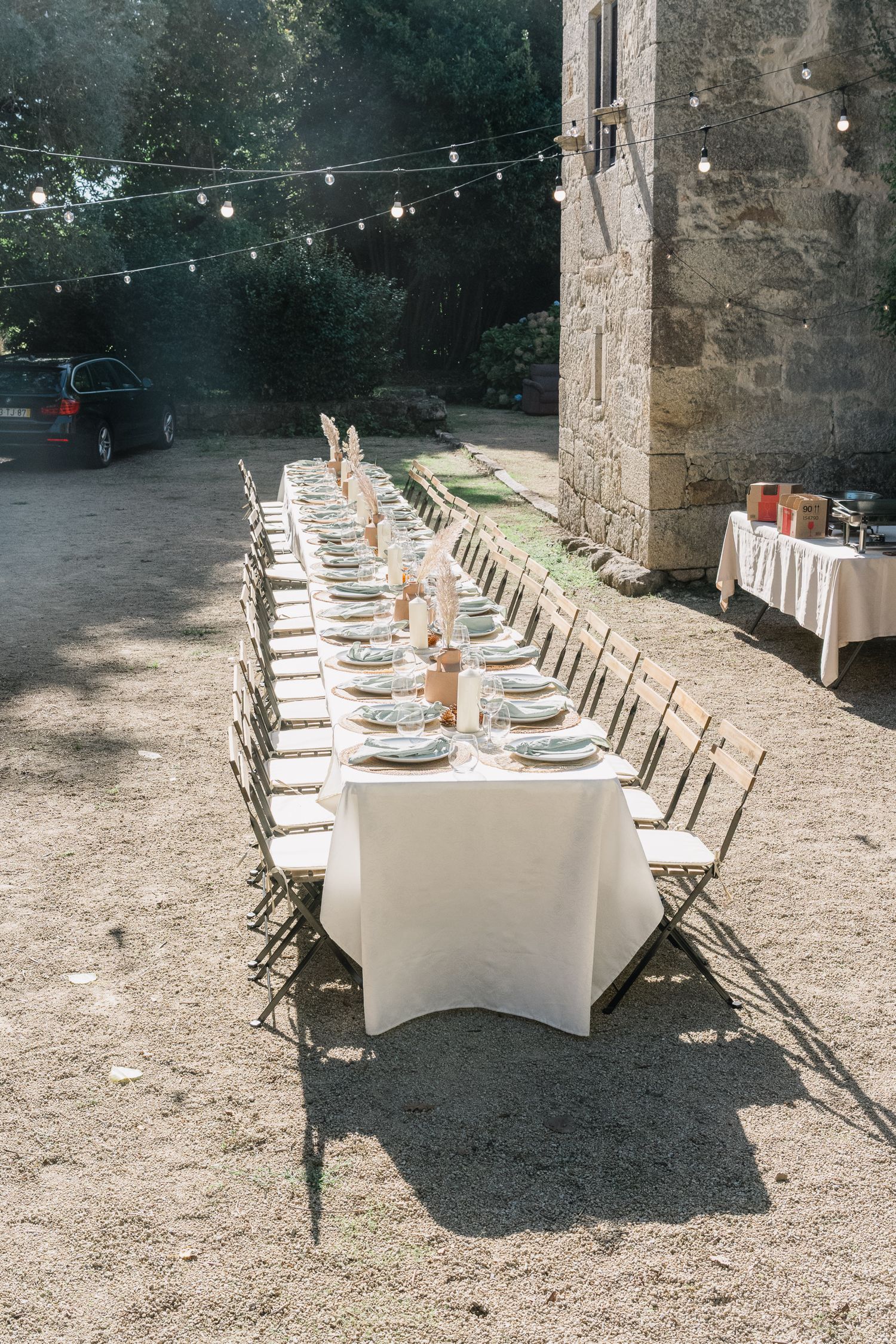Long outdoor dining table with white linens and rustic chairs set against stone wall under string lights for elegant event.