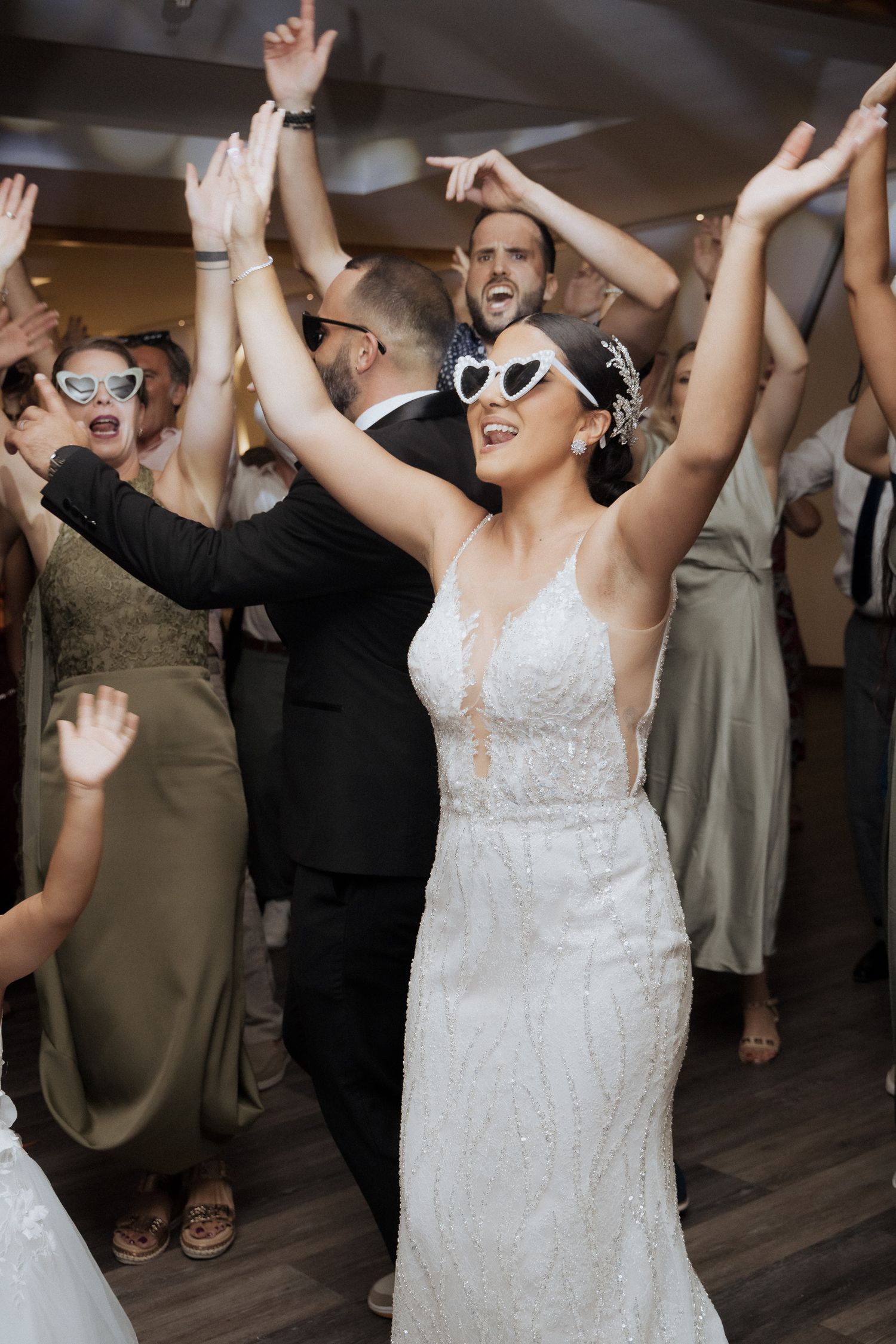 Bride and wedding party dancing joyfully with raised arms on wooden dance floor at reception celebration.
