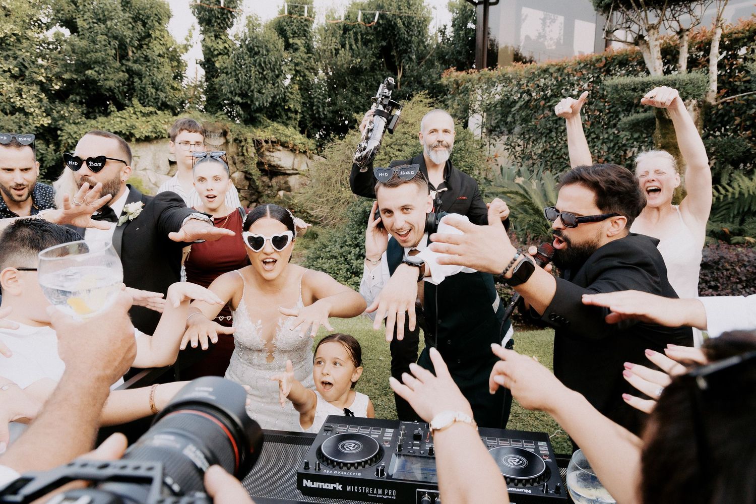 People celebrate outdoors at a party with hands raised in the air around a DJ setup in a backyard setting.