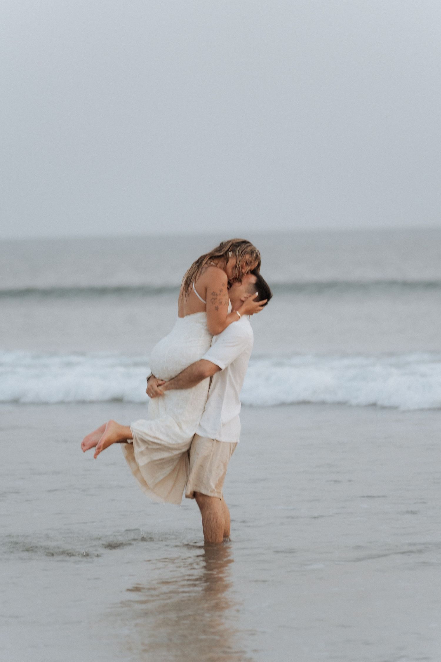 A romantic couple embraces while standing in shallow ocean waves wearing light colored clothing at the beach.