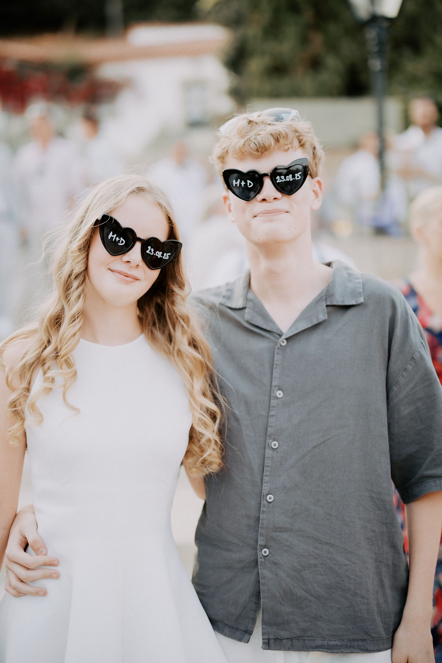 A couple wearing matching black sunglasses pose together outdoors, with one in white and one in gray attire.
