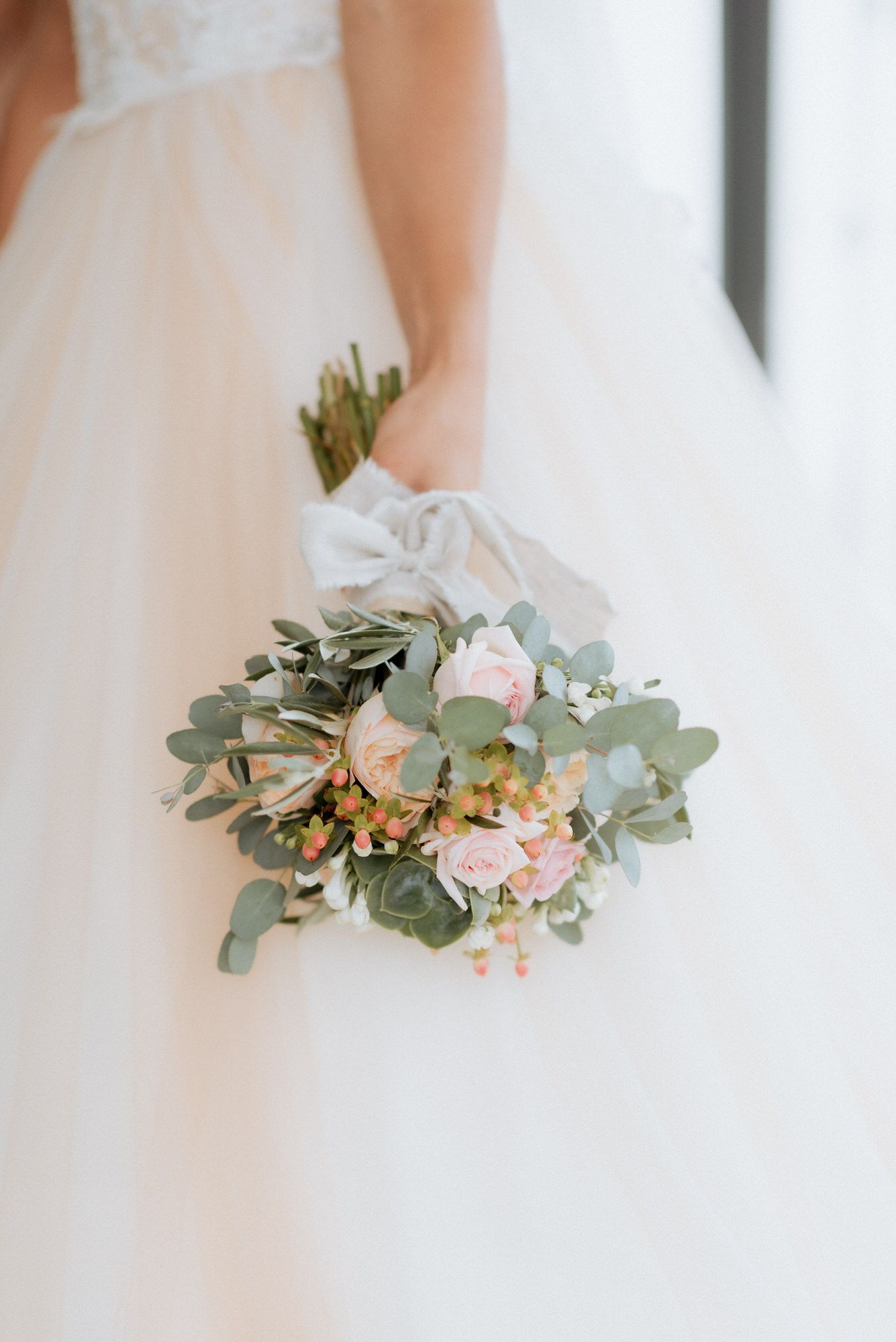 A delicate bridal bouquet featuring pale pink roses, eucalyptus leaves and small peach flowers against a white wedding dress.