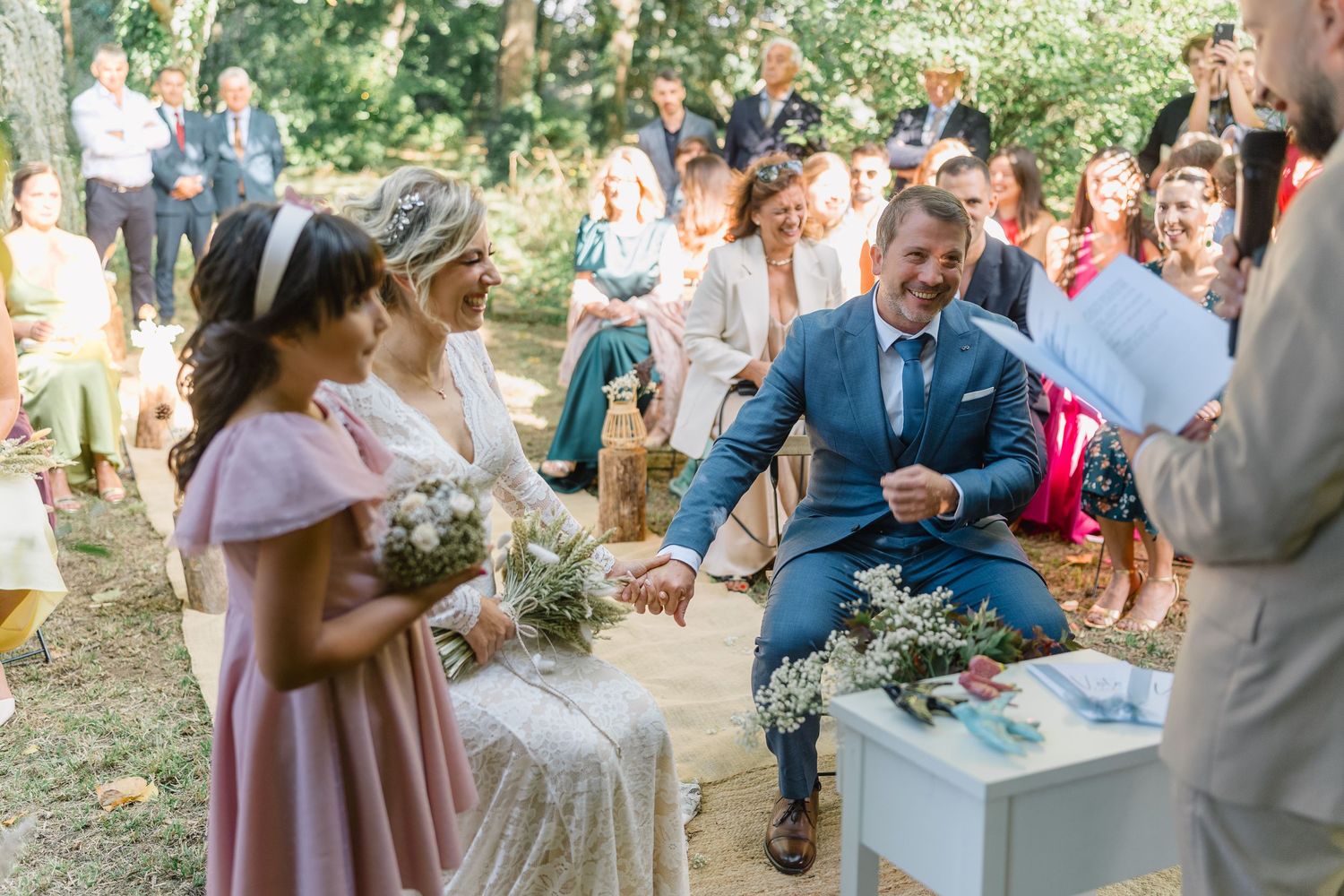 A romantic outdoor wedding celebration with guests gathered in a garden setting under trees and string lights.