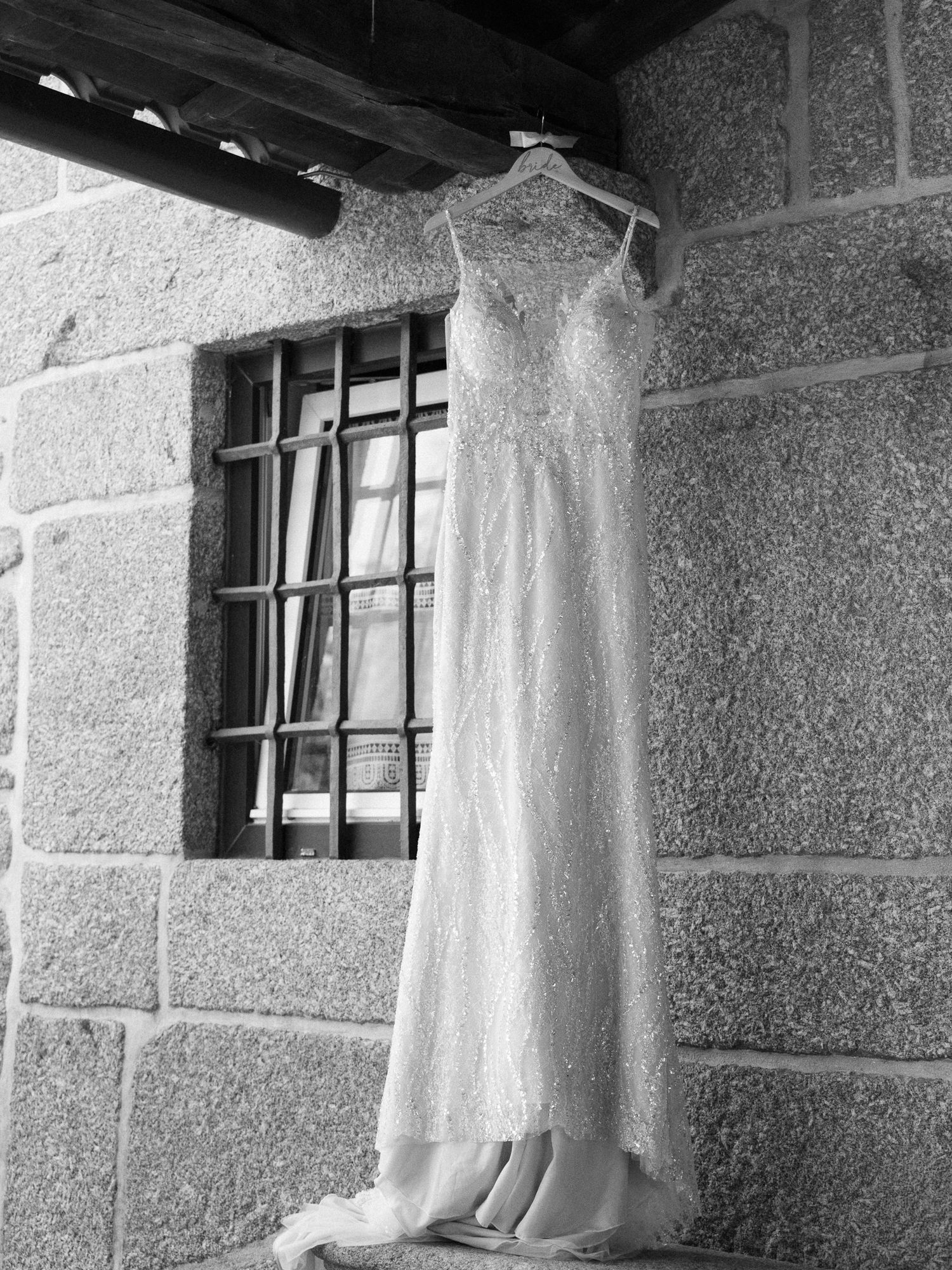 A delicate white lace wedding dress hangs on a hanger against a stone wall with a barred window in black and white.