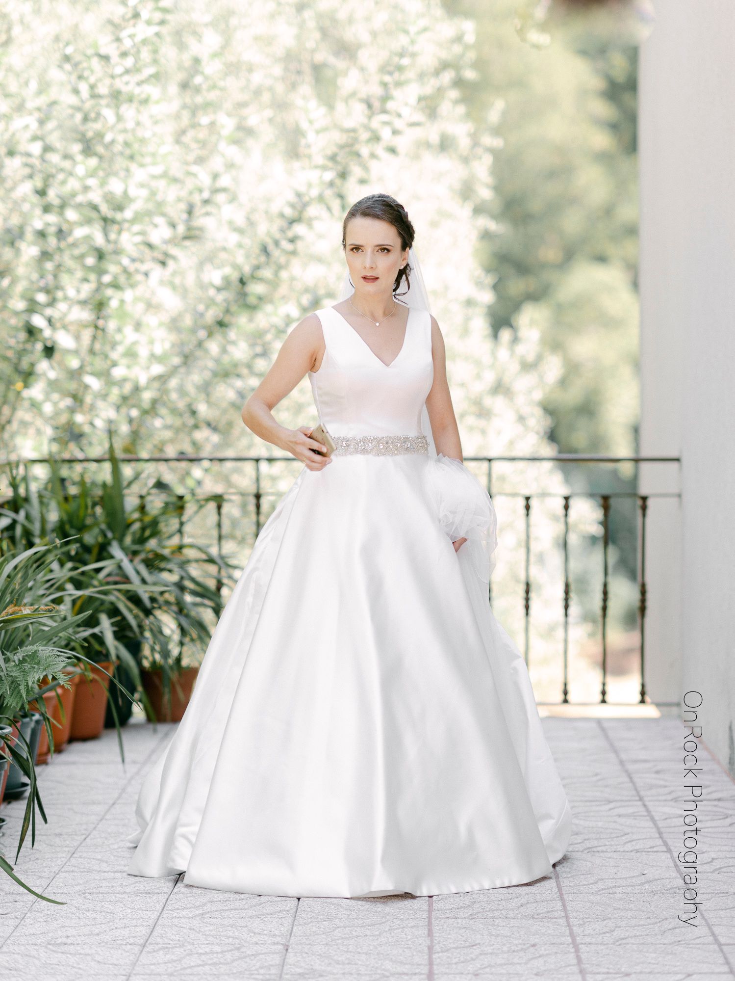 A bride in a classic white ball gown poses gracefully on an outdoor patio with white flowering trees in the background.