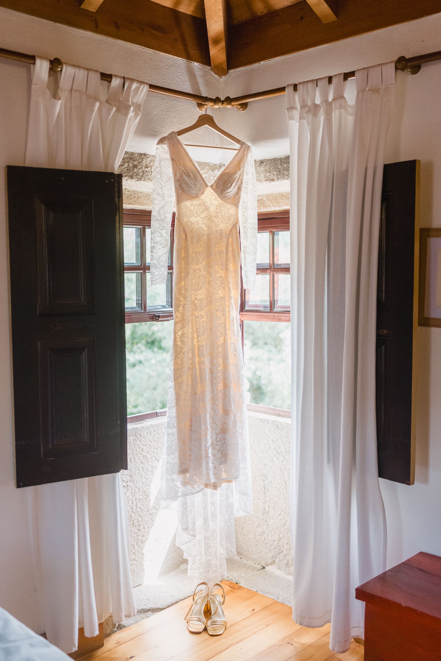 A white wedding dress hangs elegantly from wooden beams in a rustic room with white curtains and natural lighting.