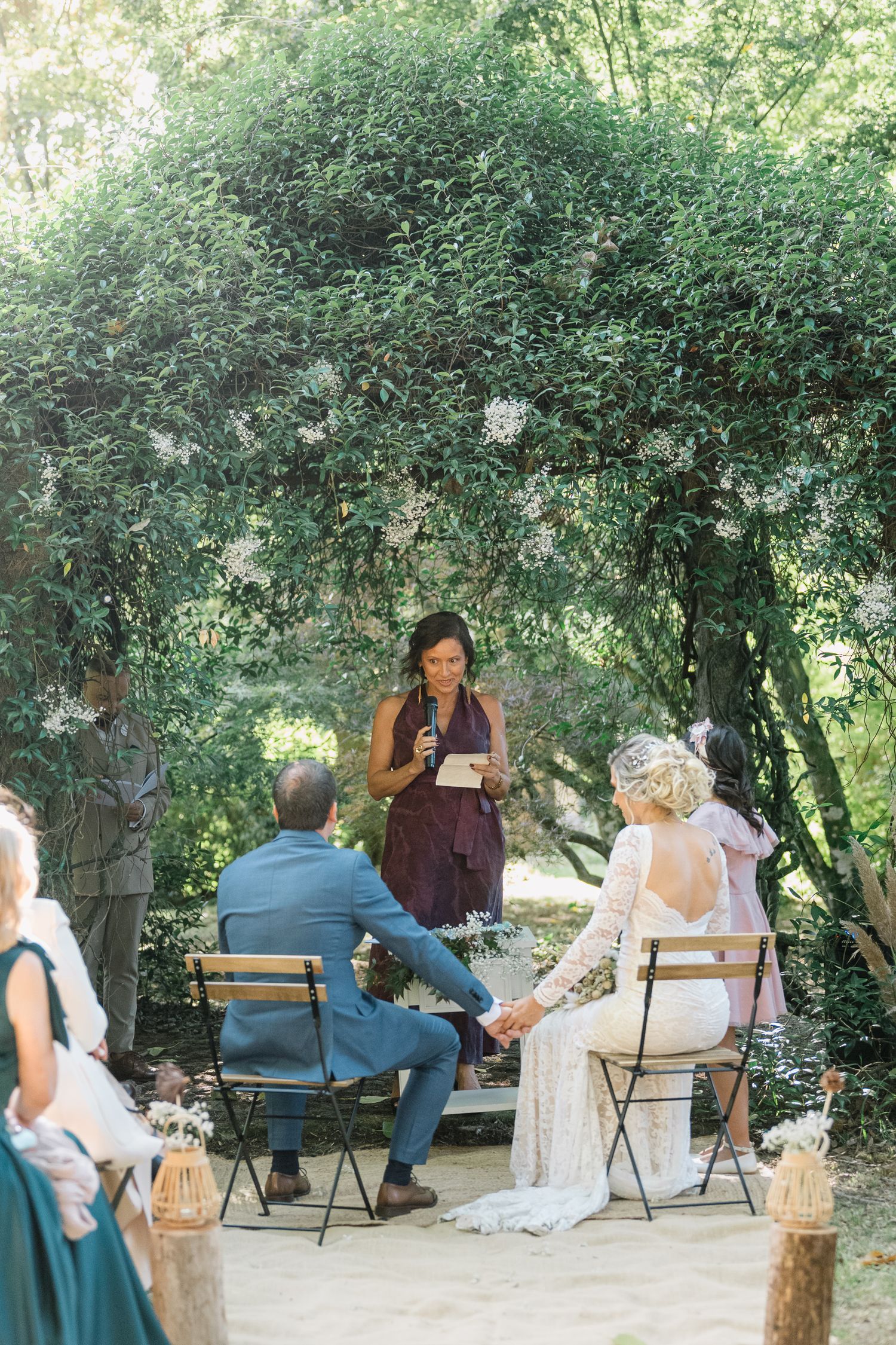 A group of people sit in folding chairs under shady trees during an outdoor gathering.