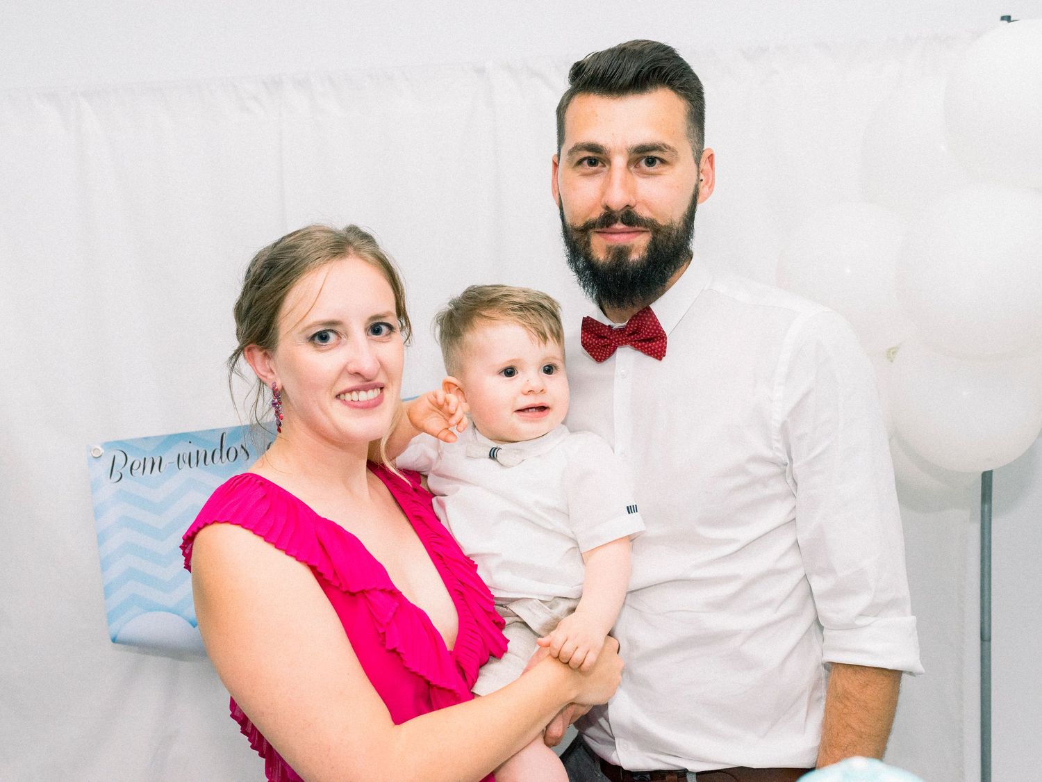 A family poses together in coordinated white and pink outfits for a studio portrait session with balloons.