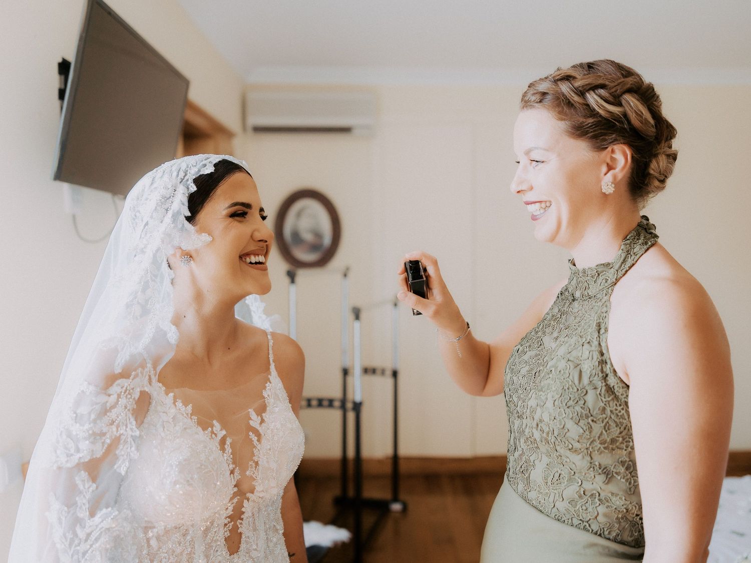Two women share a joyful moment together during wedding preparations, one wearing a white lace gown with veil.