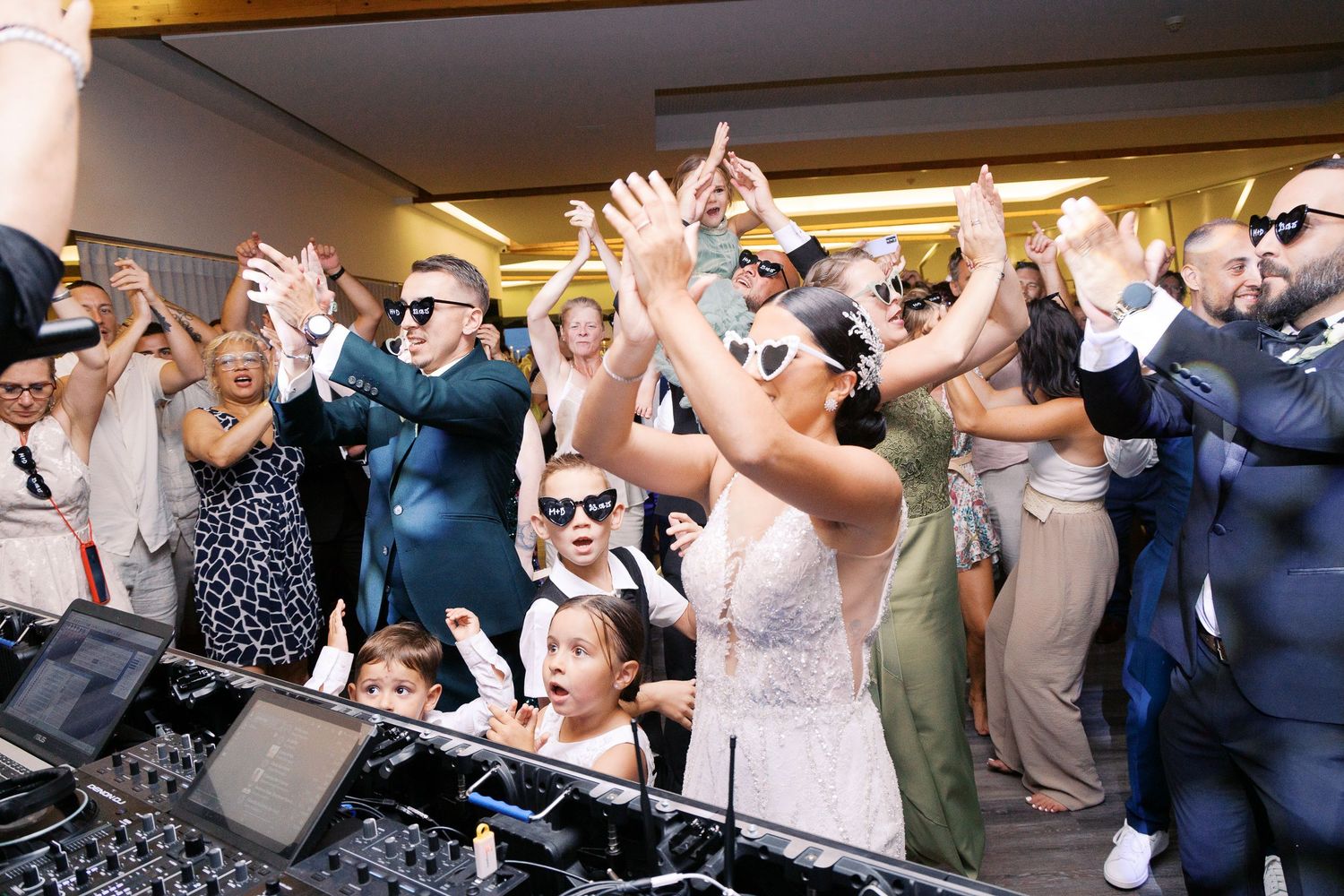 Wedding guests dancing and cheering with hands raised at a reception party while DJ plays music.