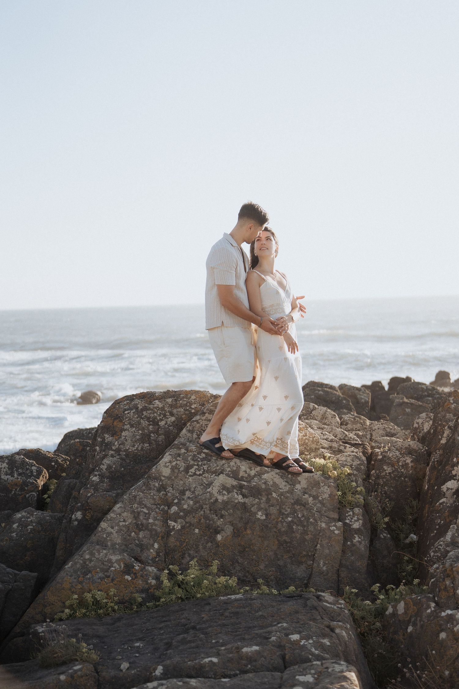 Um casal vestido de branco está sentado em clima de intimidade sobre rochas costeiras com vista para o oceano.