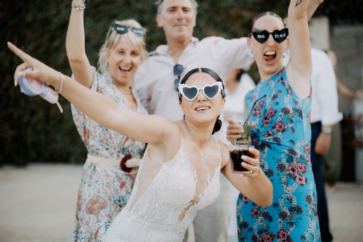 A group of wedding guests wearing sunglasses and floral dresses dance joyfully outdoors during a celebration.