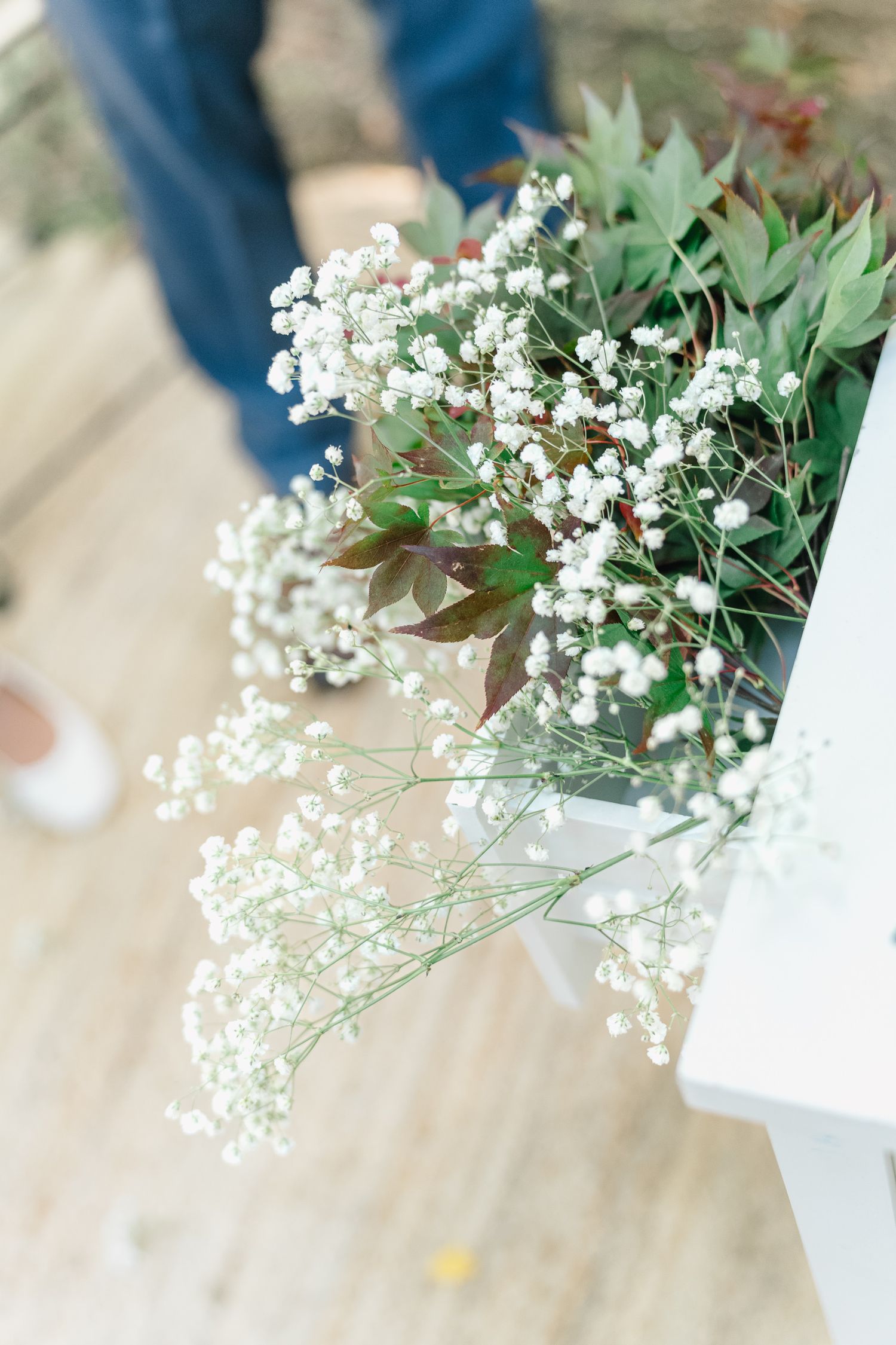 Delicate white baby's breath flowers and green foliage arranged in a white vase on a light wooden surface.