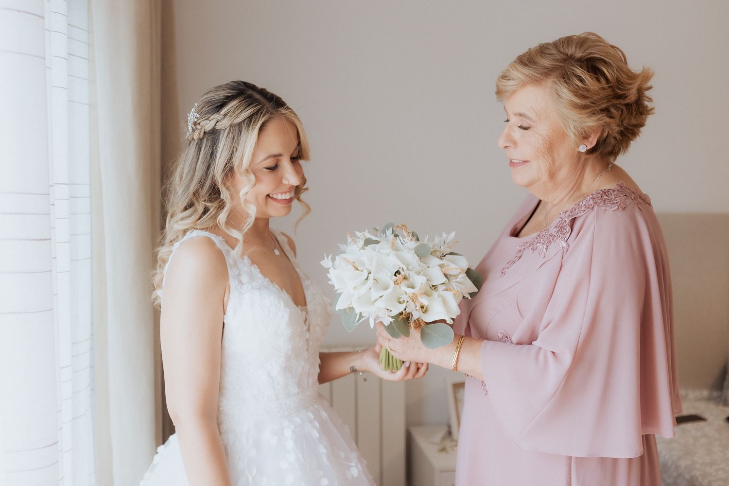 A bride in a white wedding gown shares a tender moment with someone wearing a pink dress in a sunlit room.