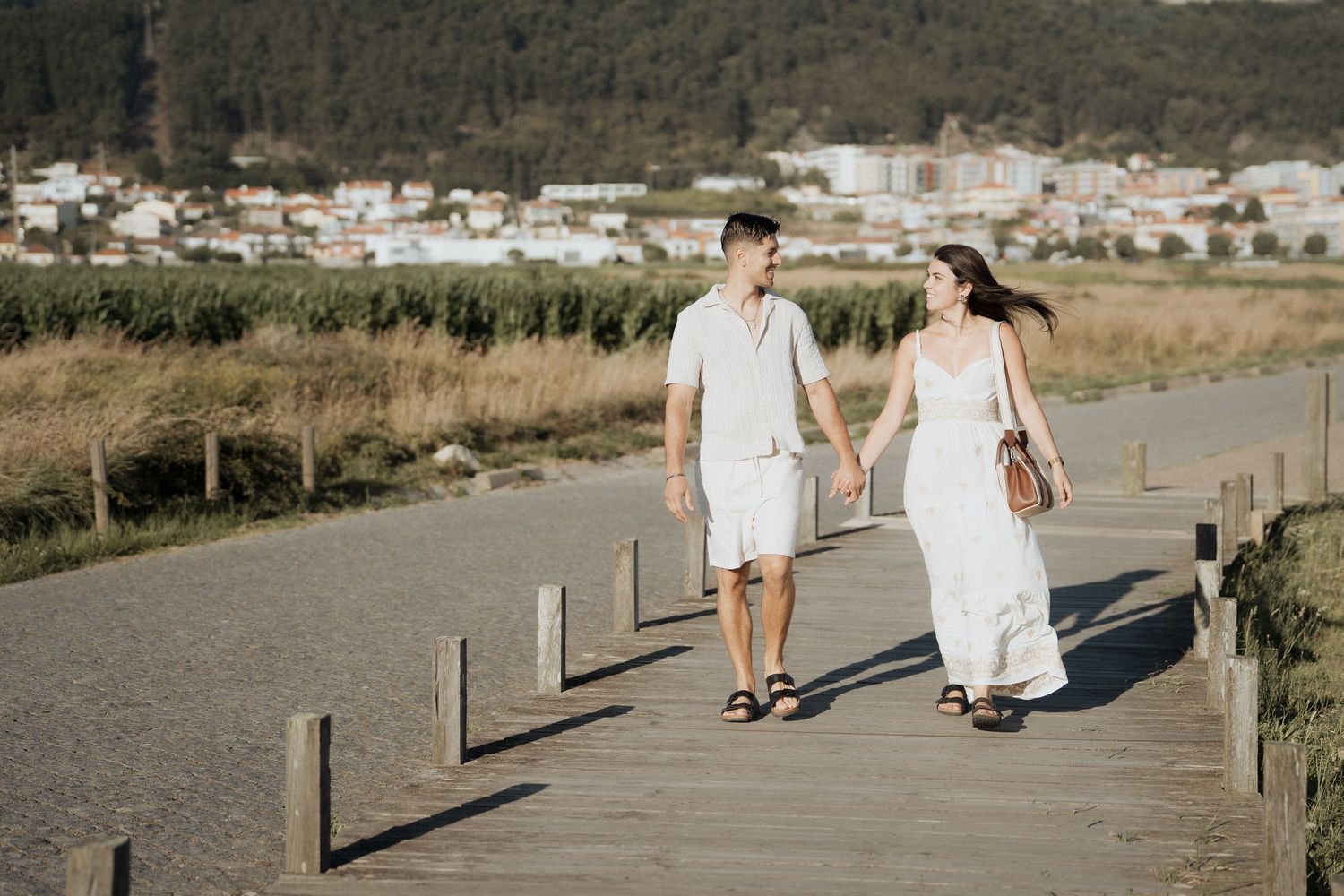 Um casal caminha de mãos dadas ao longo de um calçadão à beira-mar, com Viana do Castelo ao fundo.