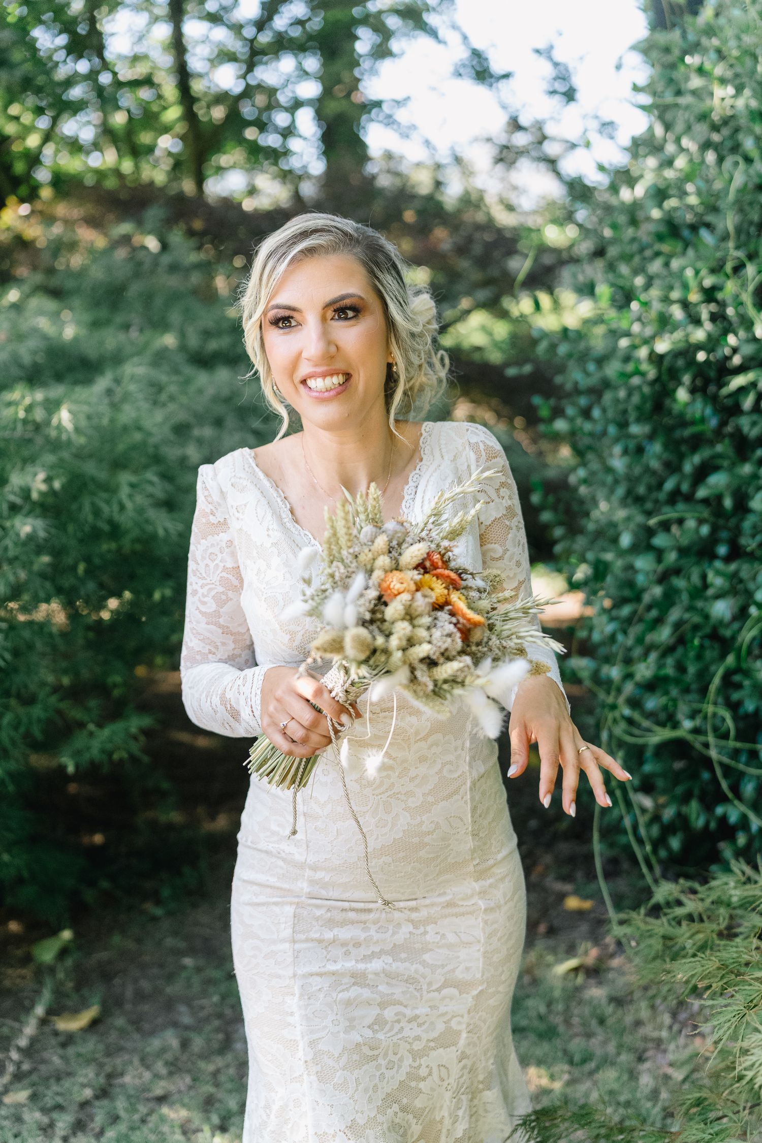 Noiva com vestido de renda exibe o seu anel e o bouquet de flores secas num jardim ao ar livre. Casamento em ponte de lima