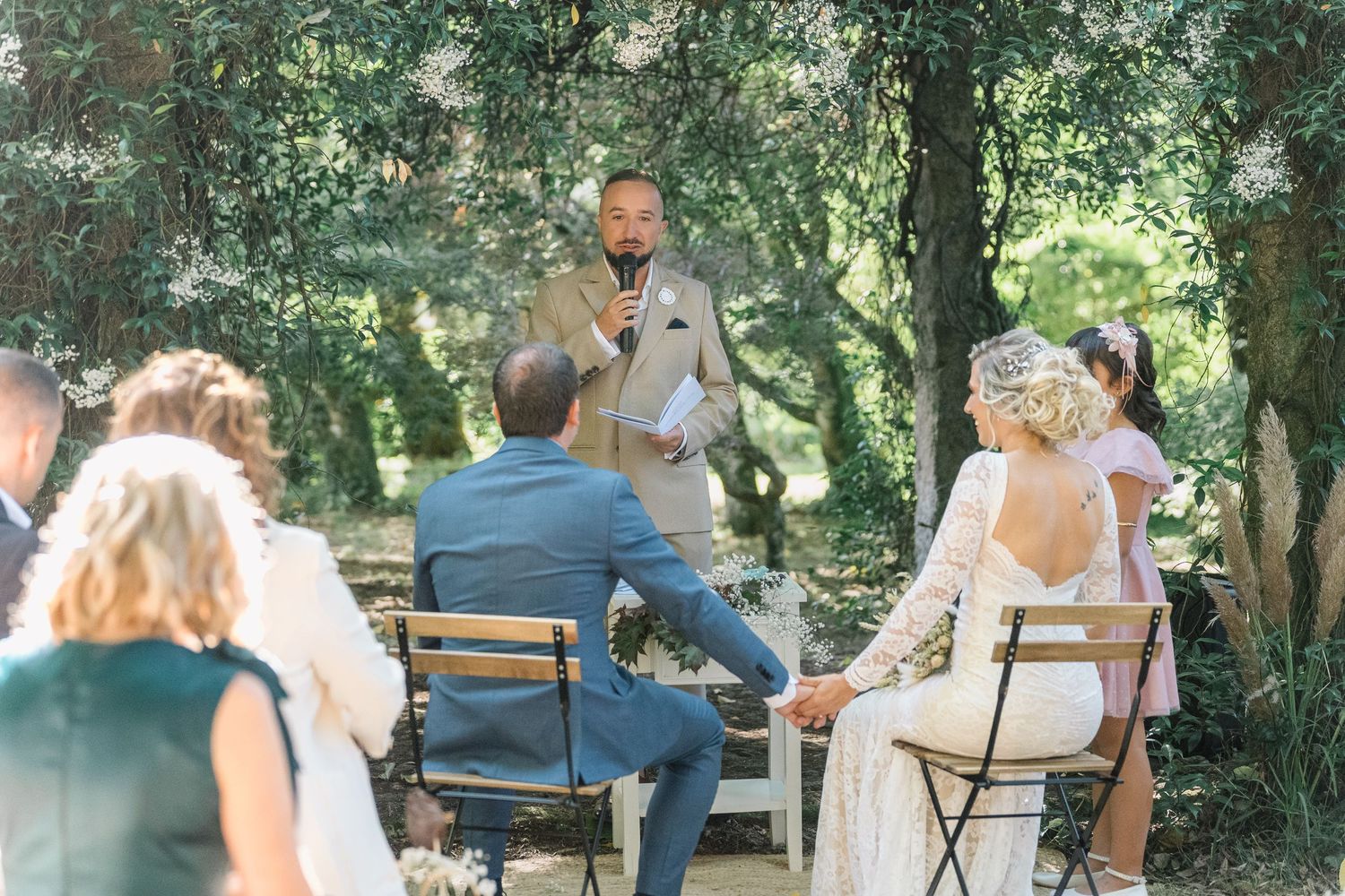 People gather outdoors for a casual celebration in a wooded garden setting with chairs and tables arranged under trees.