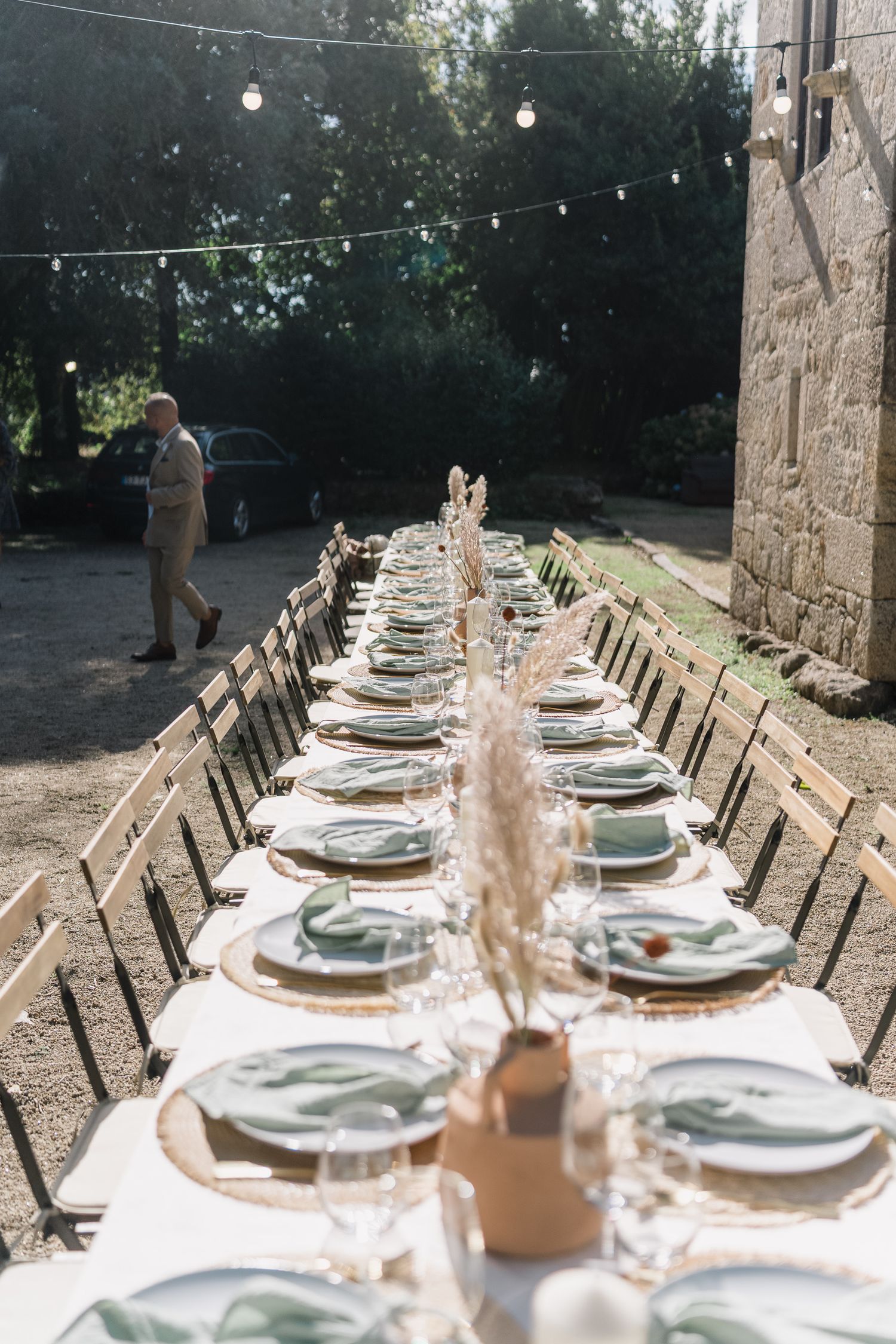 Long rustic outdoor dining table set with white linens and string lights overhead for an elegant garden party.