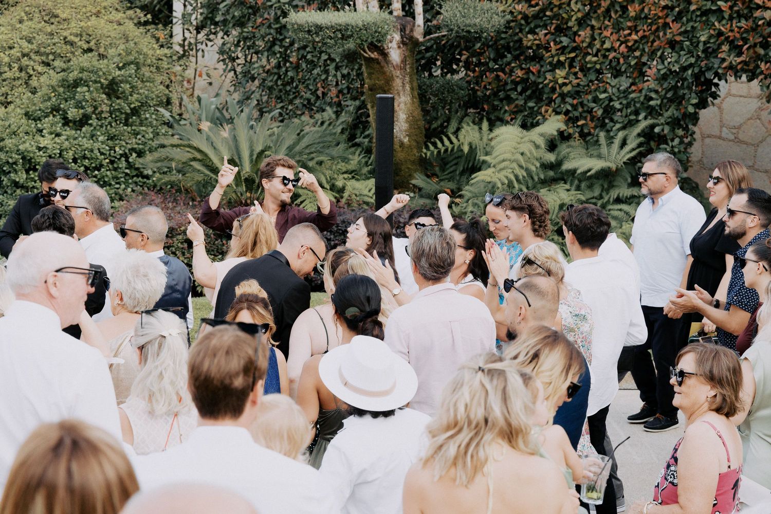 A crowd of guests dressed in white gather outdoors for a summer garden celebration.