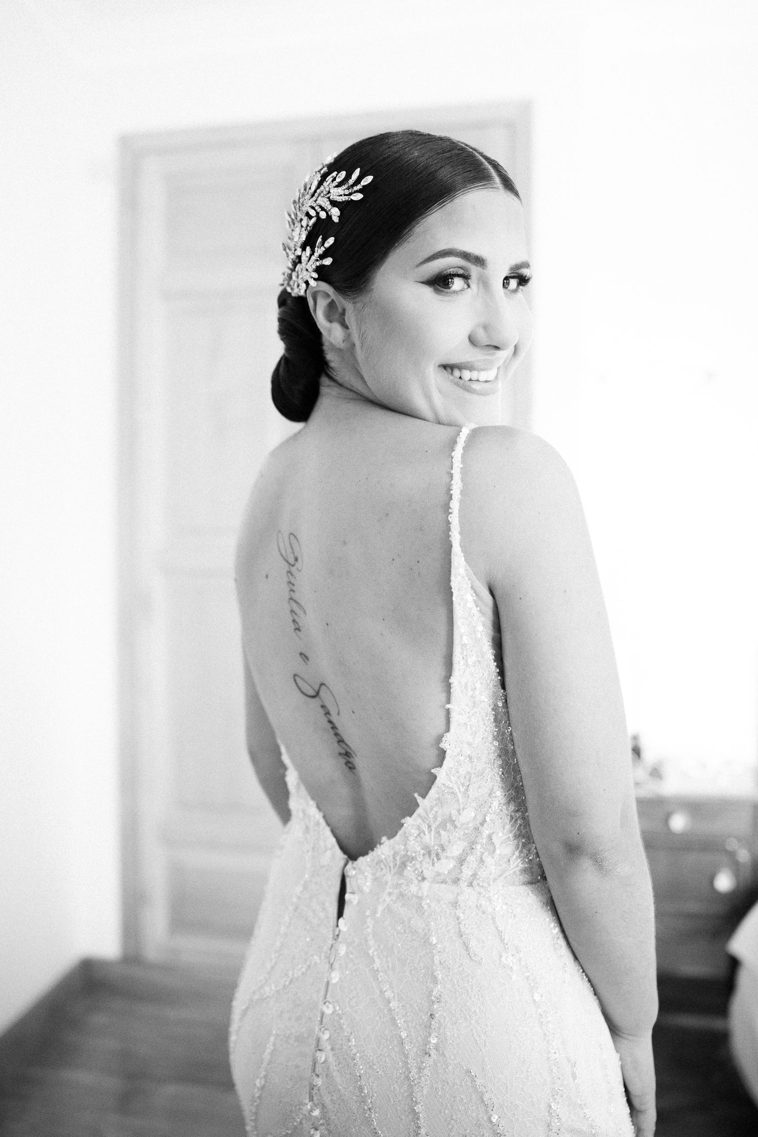 Black and white portrait of a bride in a backless lace wedding dress with a hair accessory looking over her shoulder.