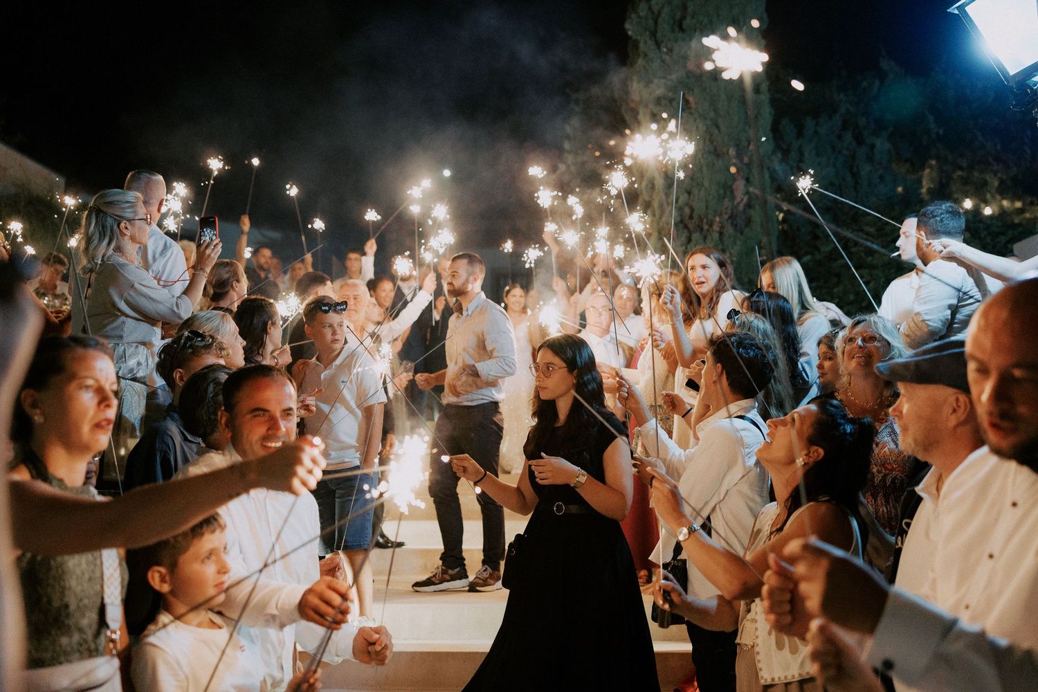 Wedding guests hold sparklers creating a magical nighttime tunnel effect at an outdoor celebration.