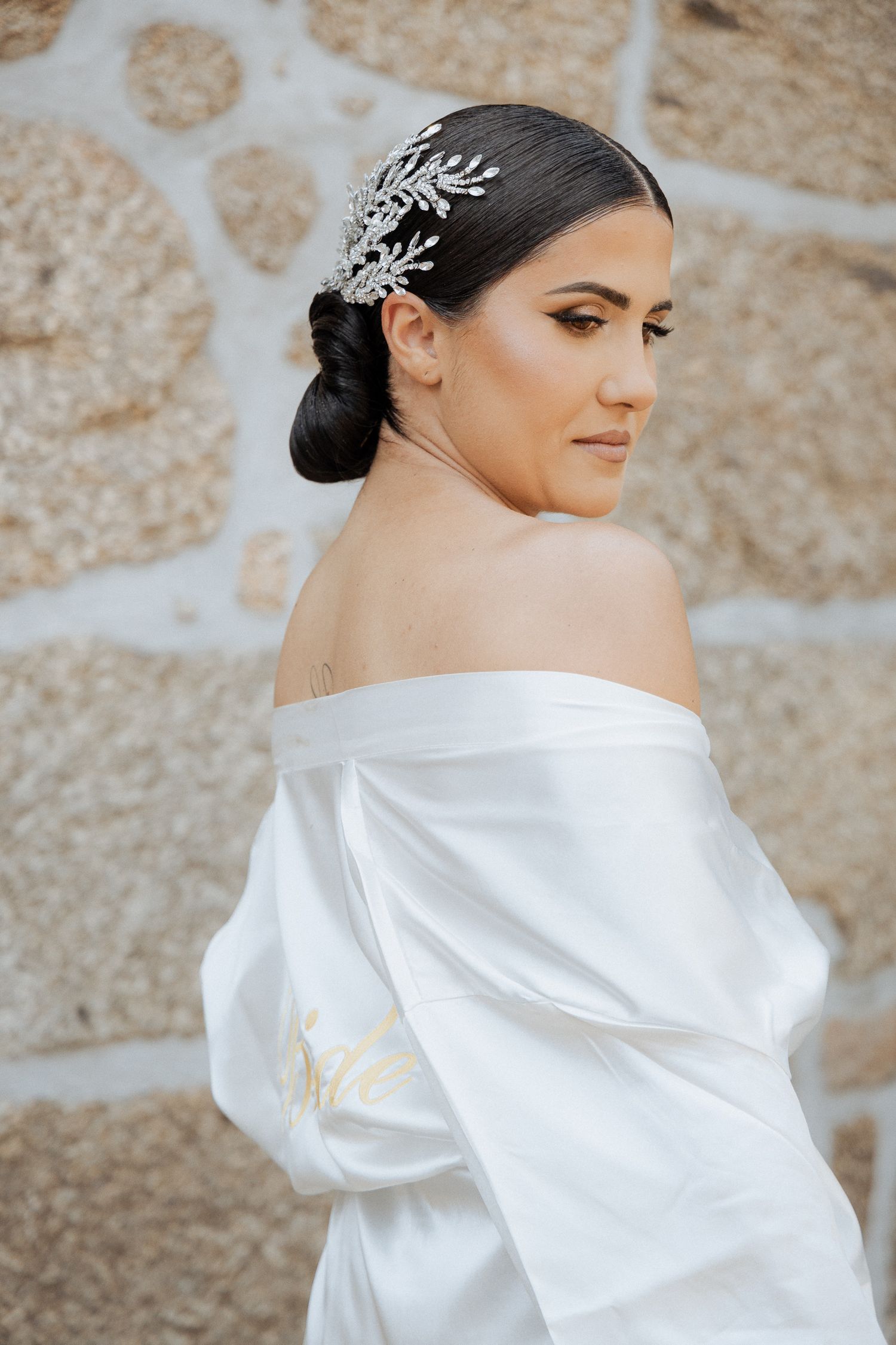 Bride wearing white satin robe and crystal hair accessory poses against stone wall in elegant wedding preparation photos.