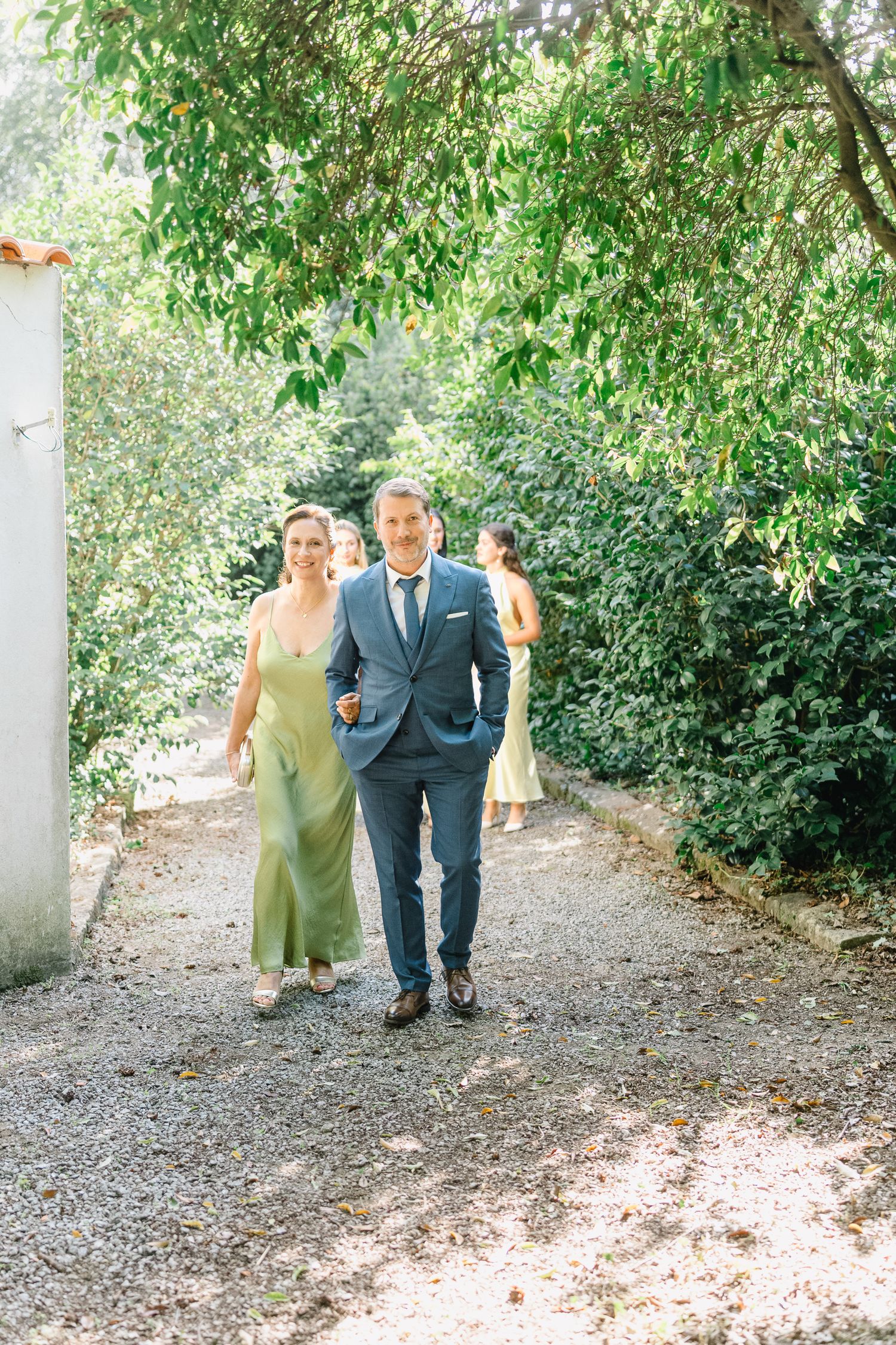 Wedding party members walk together down a sunlit garden path surrounded by lush green foliage.