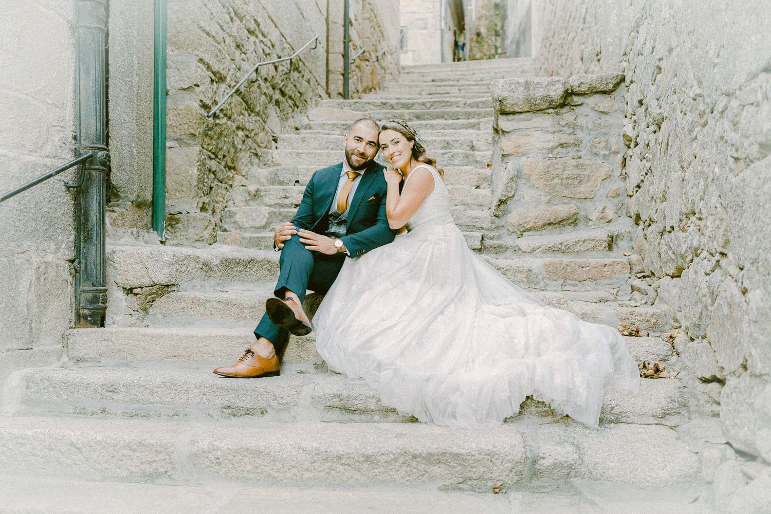 A couple in wedding attire sits together on stone steps, with the bride in white and groom in emerald green suit.