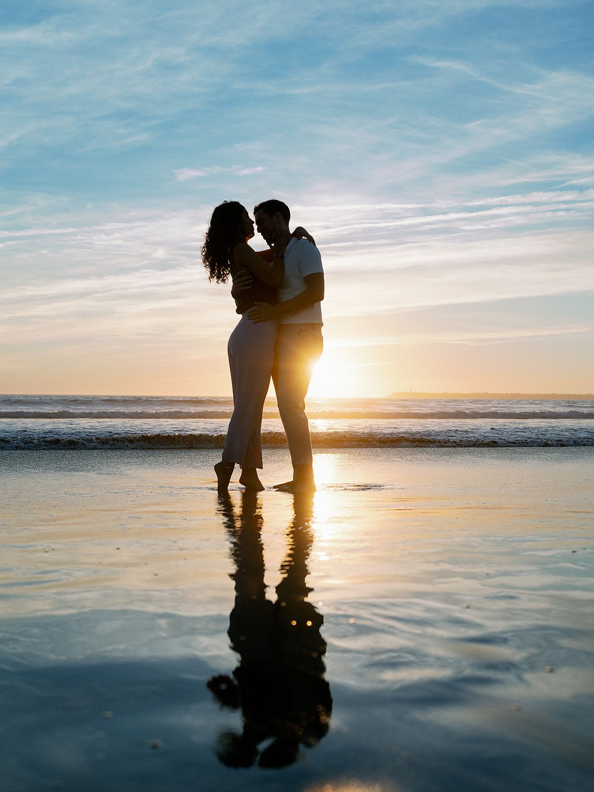 Silhouetted couple standing in shallow reflective water at sunset creates romantic beach scene with glowing sky.