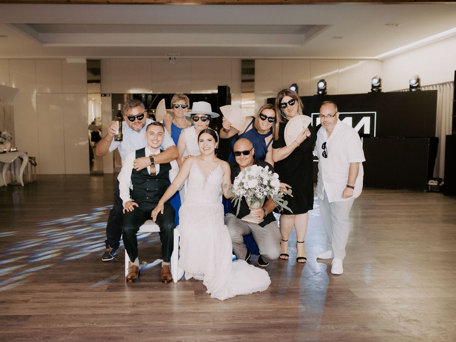 Wedding party poses together on dance floor at indoor reception venue with decorative lighting.