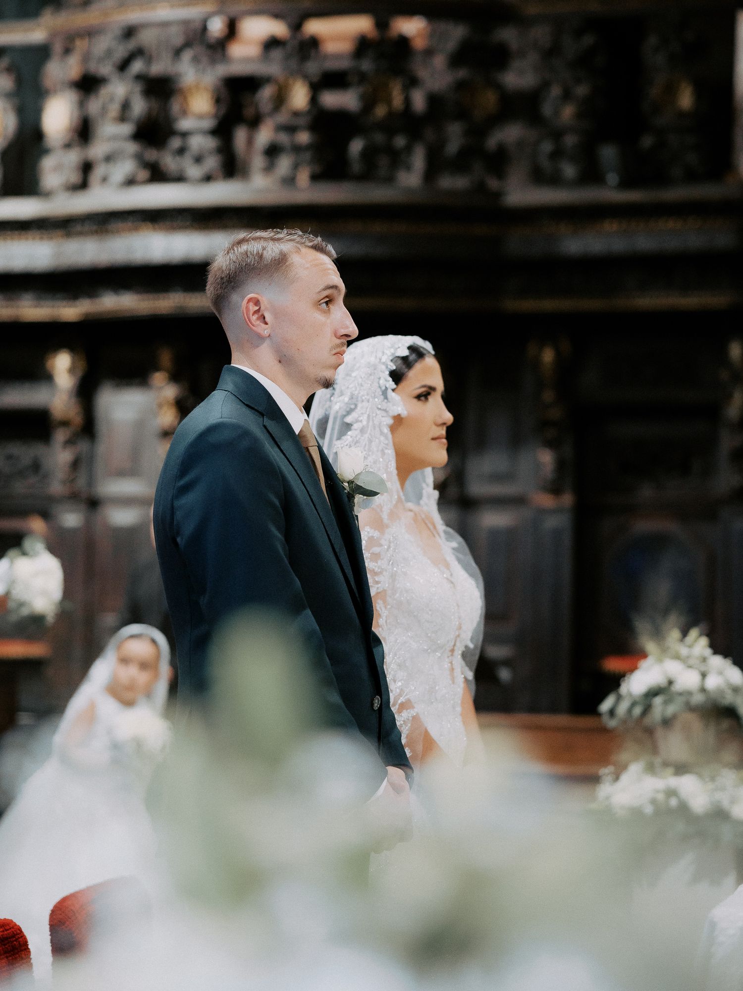 A couple stands together during their wedding ceremony in a traditional church with ornate wooden details.