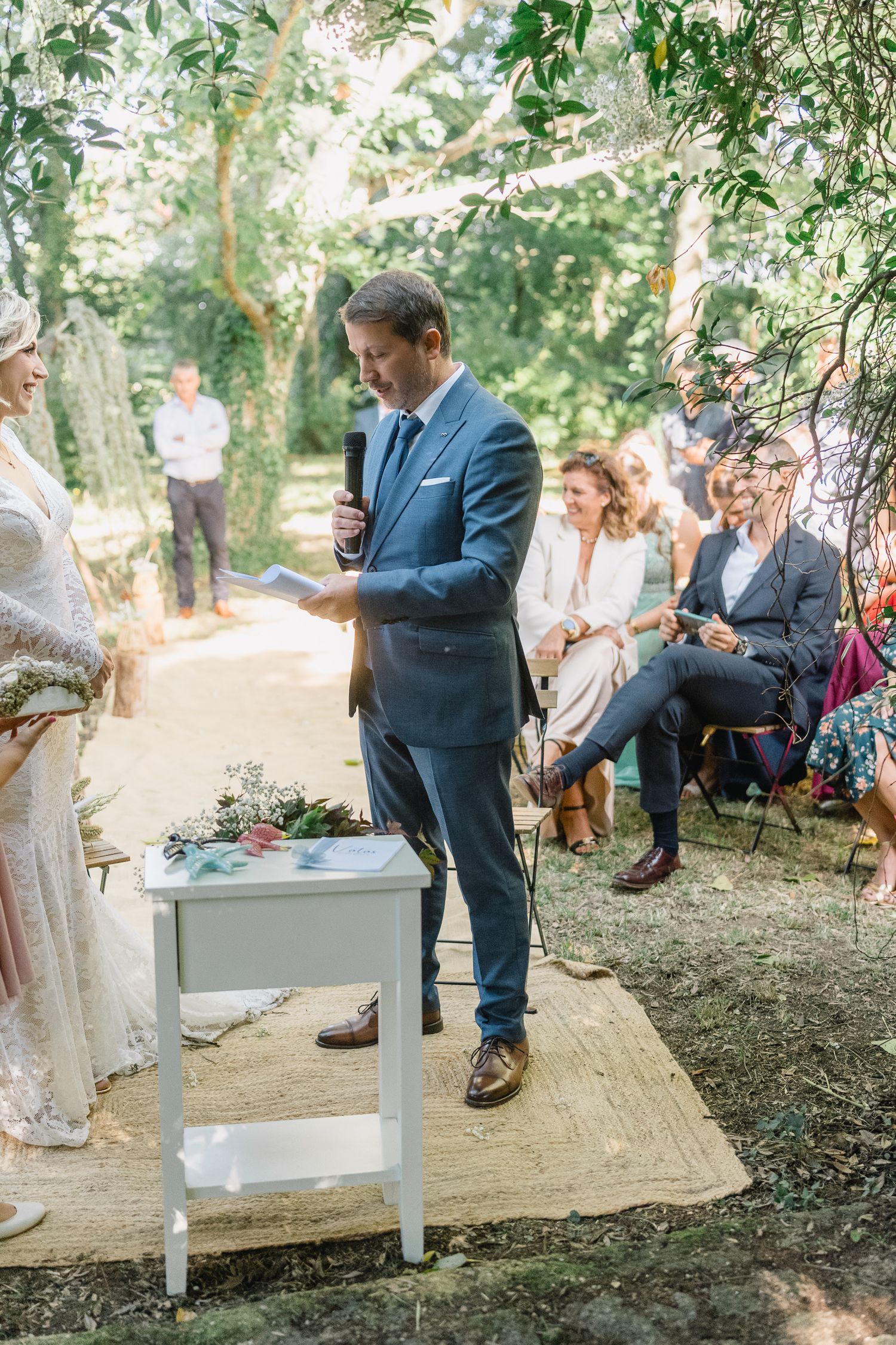 An outdoor wedding ceremony in a wooded setting with rustic white benches and guests gathered under trees.