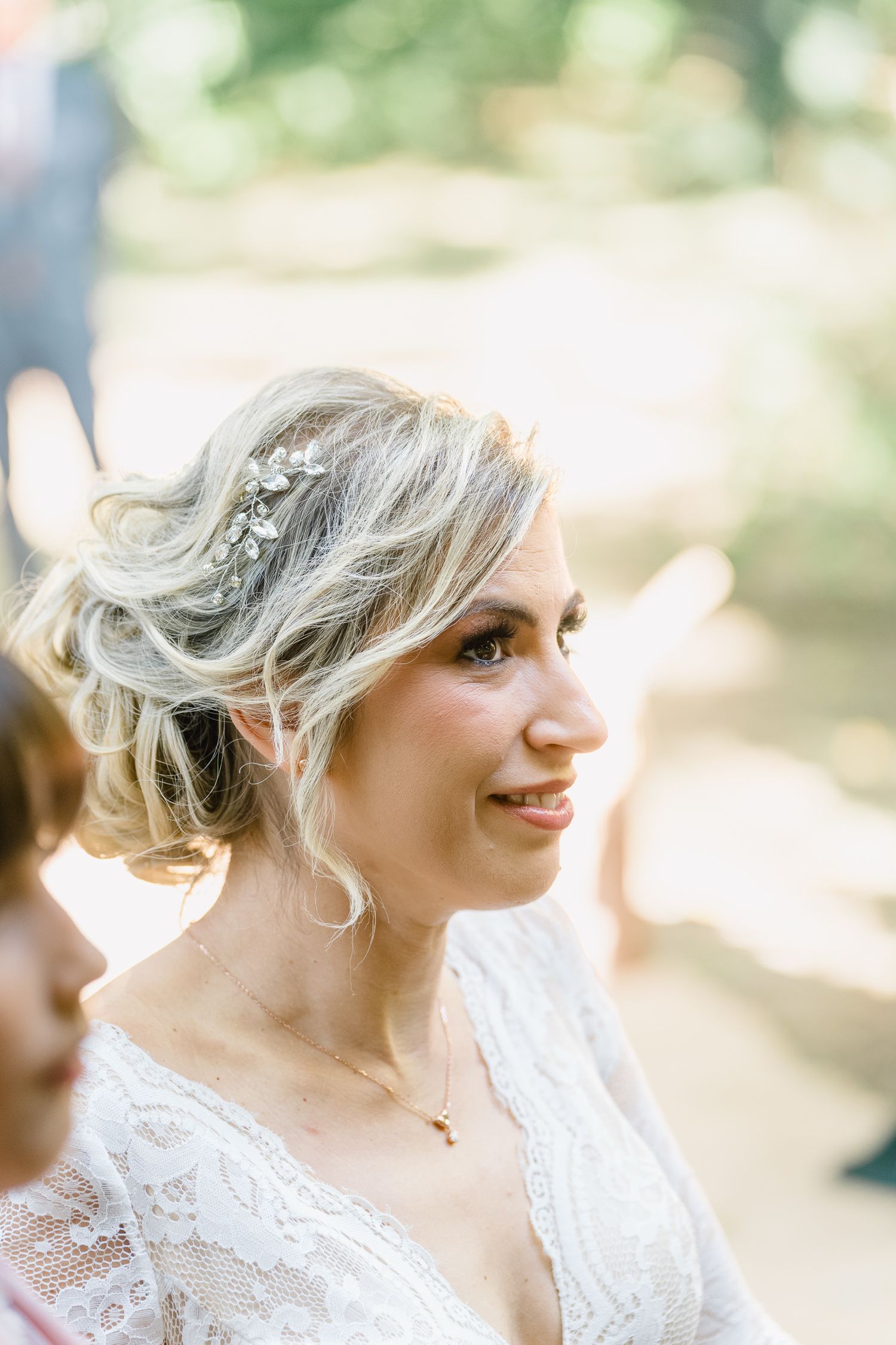 A bride in a white lace wedding dress smiles while looking to the side in a sun-dappled outdoor setting.