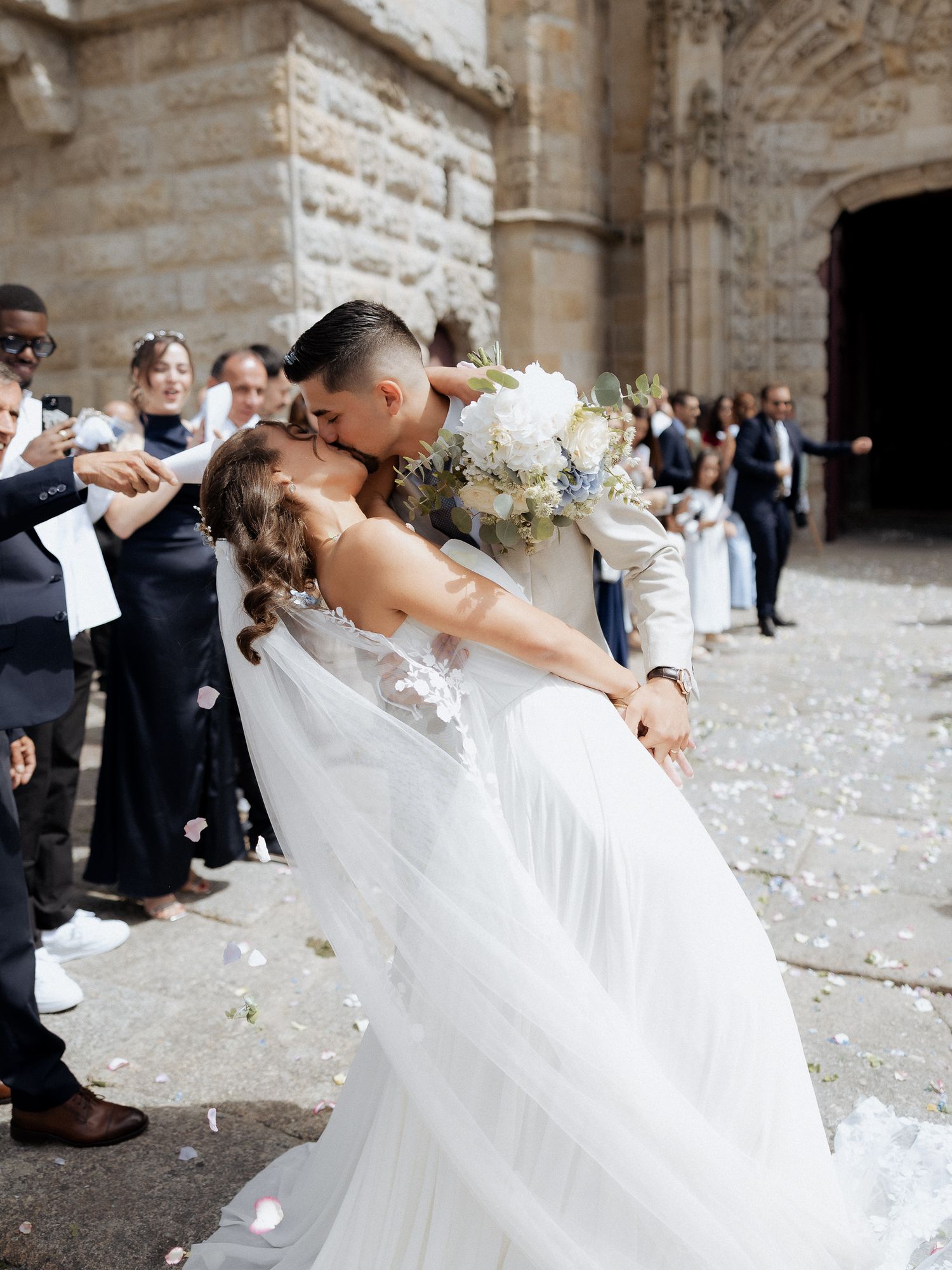 Newlywed couple sharing a romantic kiss outside a stone church while guests toss flower petals in celebration.