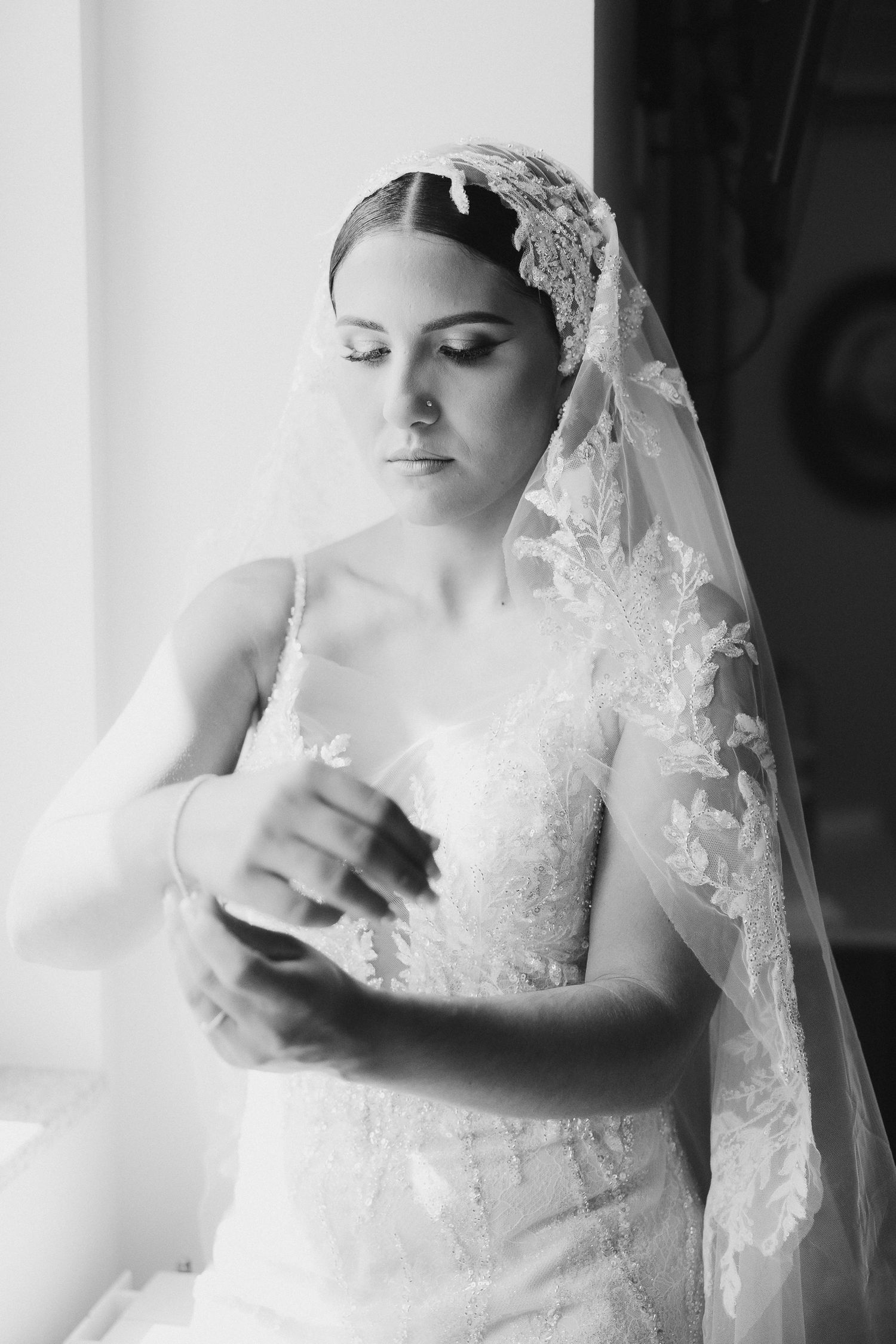 A bride in a lace wedding dress and veil stands near a window in an elegant black and white portrait.