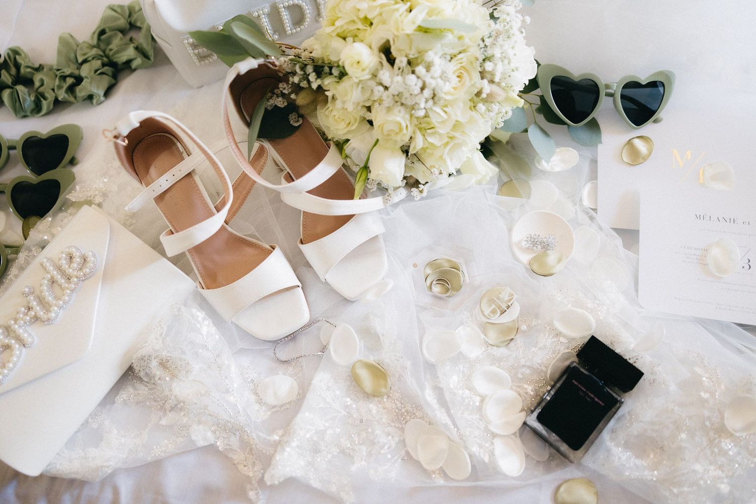 Wedding details including white flowers, sunglasses, and bridal accessories arranged on a light surface.